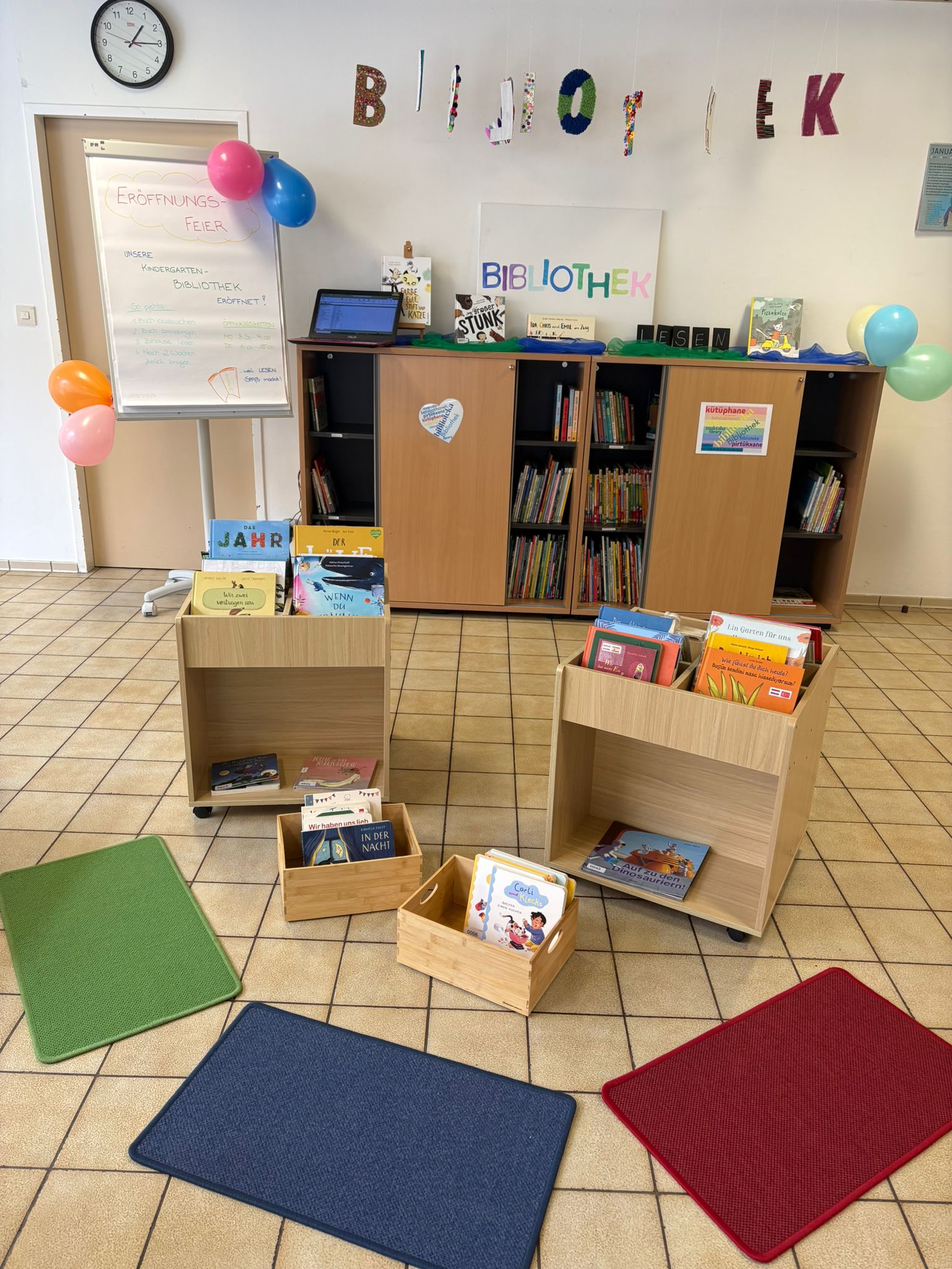 Kinderbibliothek-Ecke: Holzkisten mit Bilderbüchern, Laptop auf Schrank, bunte Ballons, farbige Teppiche auf Fliesenboden.