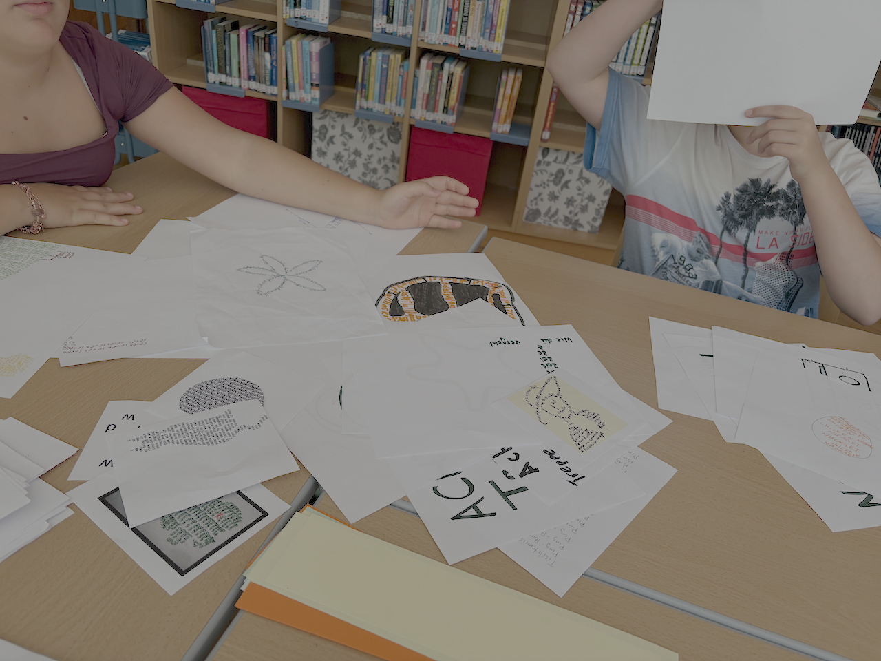 Zwei Personen arbeiten an Bastelarbeiten auf einem Bibliothekstisch; eine Person versteckt ihr Gesicht mit einem Blatt.