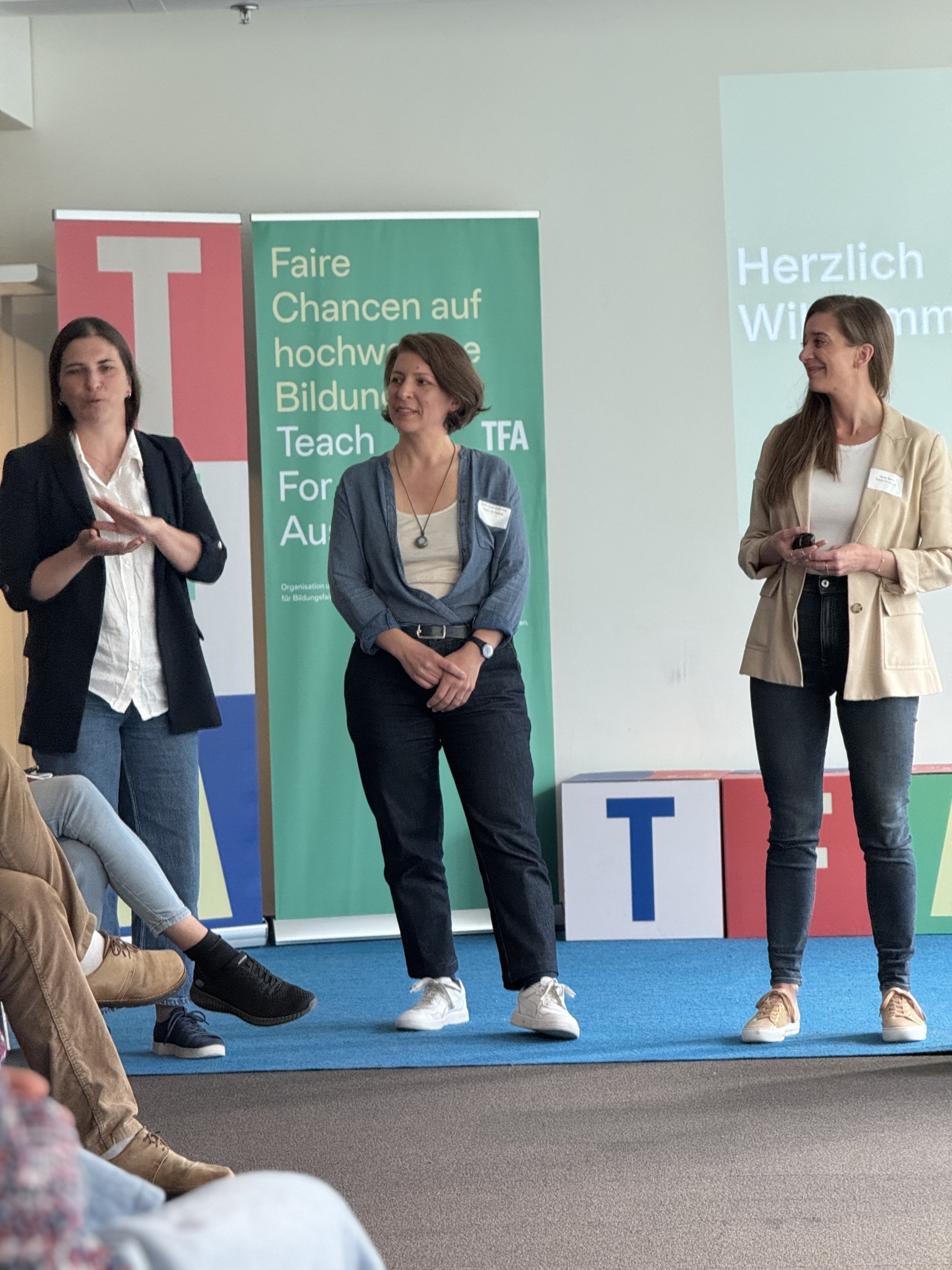 Drei Rednerinnen stehen auf einer Bühne mit blauem Teppich; grüne Banner im Hintergrund.
