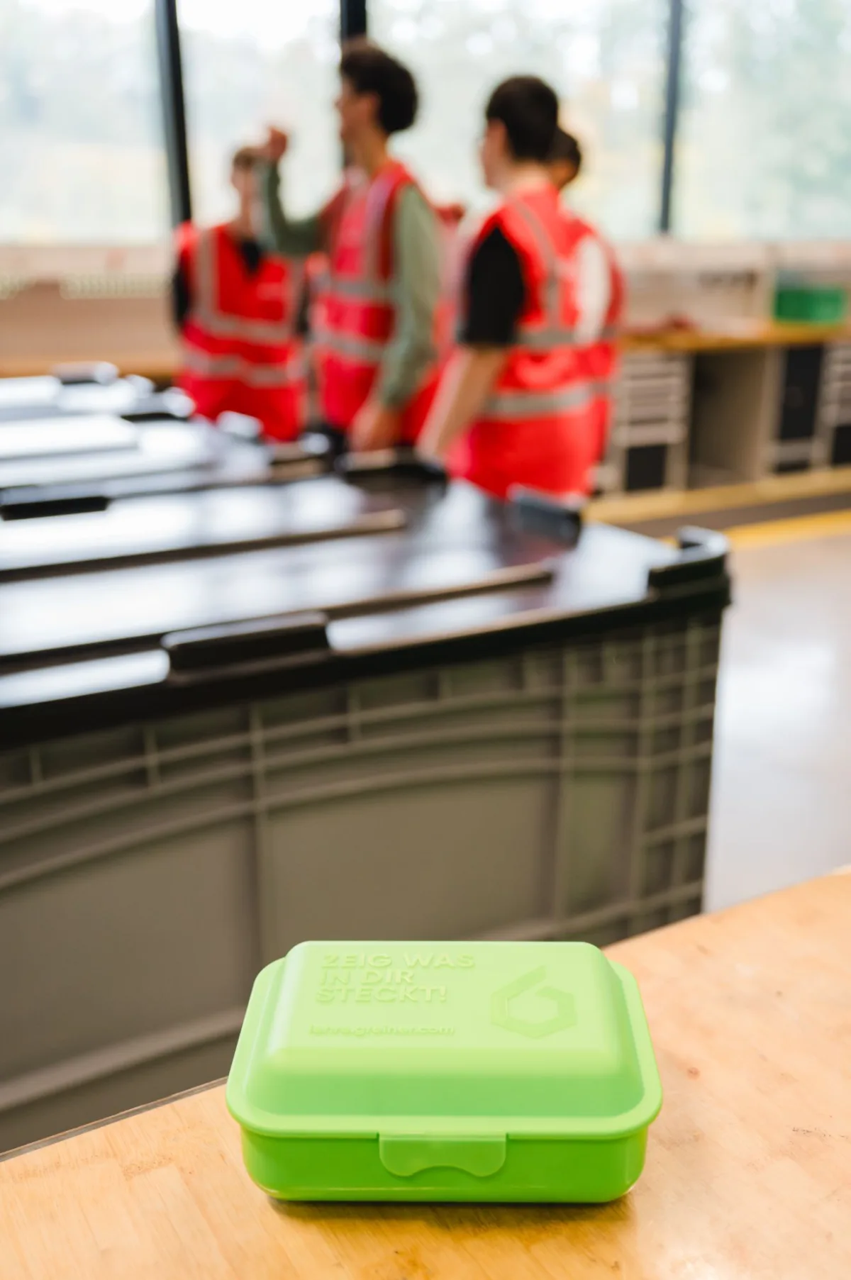 Grüne Brotdose liegt auf einem Holztisch; im Hintergrund eine Gruppe von Personen in roten Sicherheitswesten in einer Werkstatt.