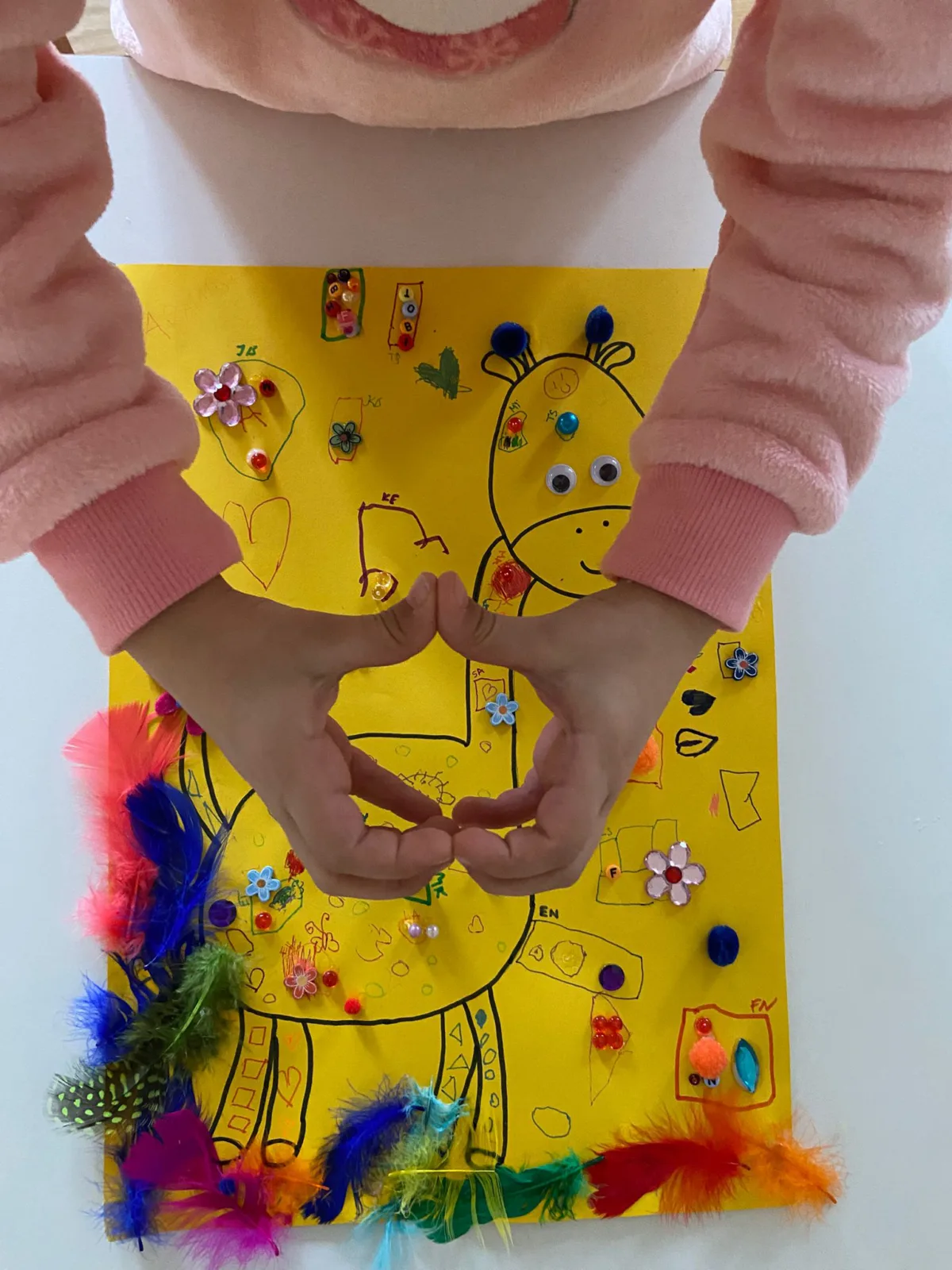 Kind formt mit den Händen ein Herz vor einem gelben Zeichenblatt mit bemalter Giraffe, verziert mit Federn und Perlen.