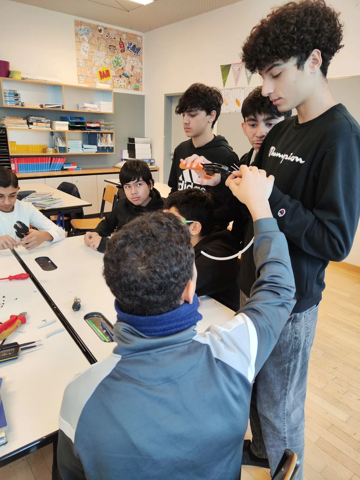 Gruppe jugendlicher Lernender im Klassenzimmer arbeitet an einem Elektronikprojekt; einer steht und hält eine Zange.