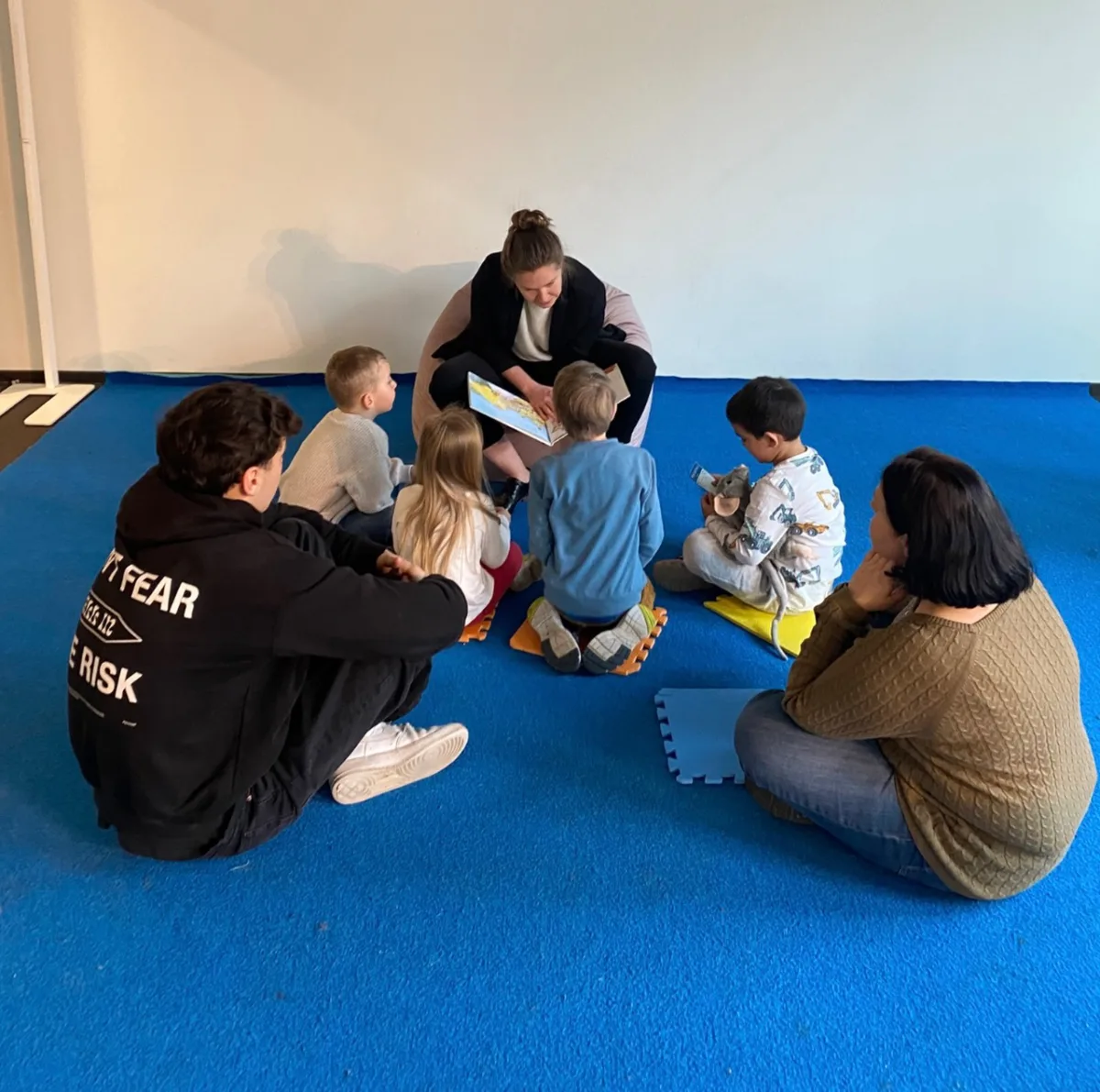 Pädagogische Fachkraft sitzt mit einer Kindergruppe im Kreis auf blauen Matten und liest ein Buch vor.