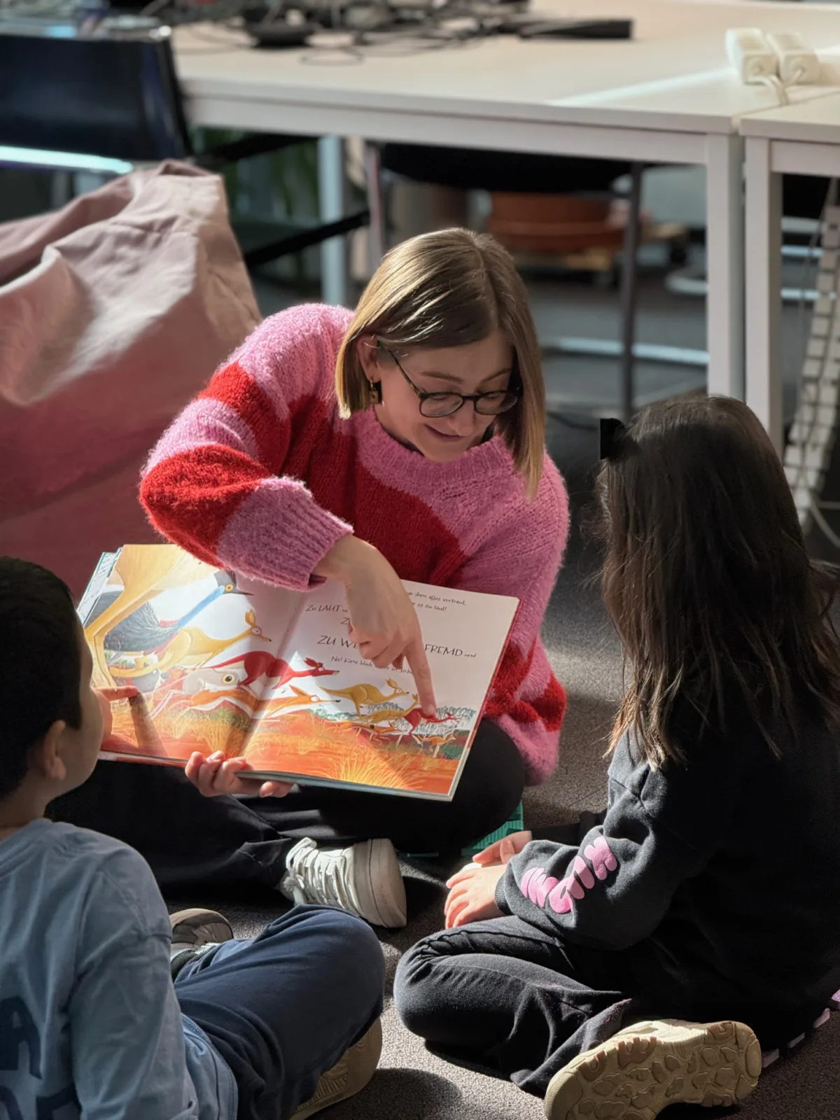 Person im pink-roten Strickpullover sitzt auf dem Boden in einem Klassenraum und liest zwei Kinder ein buntes Bilderbuch vor.