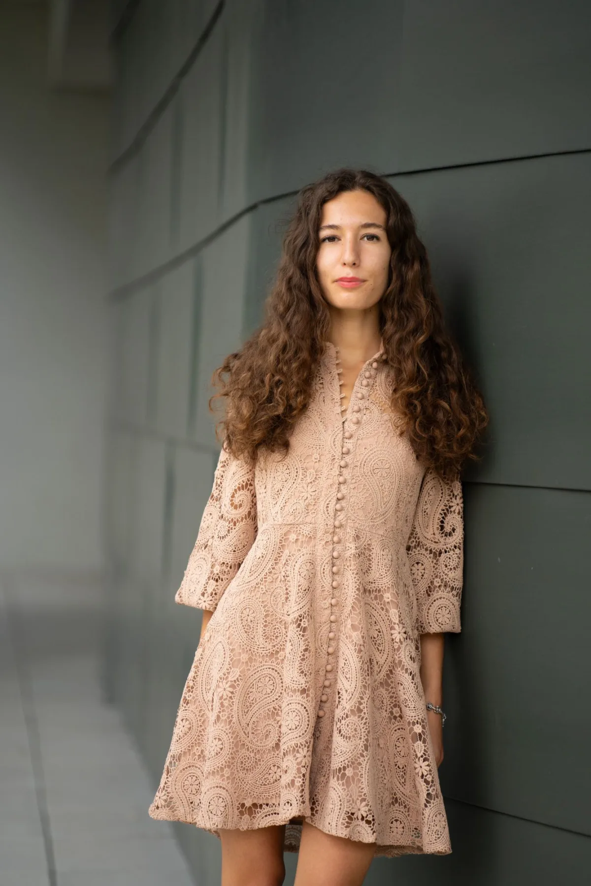 Person mit lockigen braunen Haaren trägt beiges Spitzenkleid, steht an dunkler Wand und blickt neutral in die Kamera