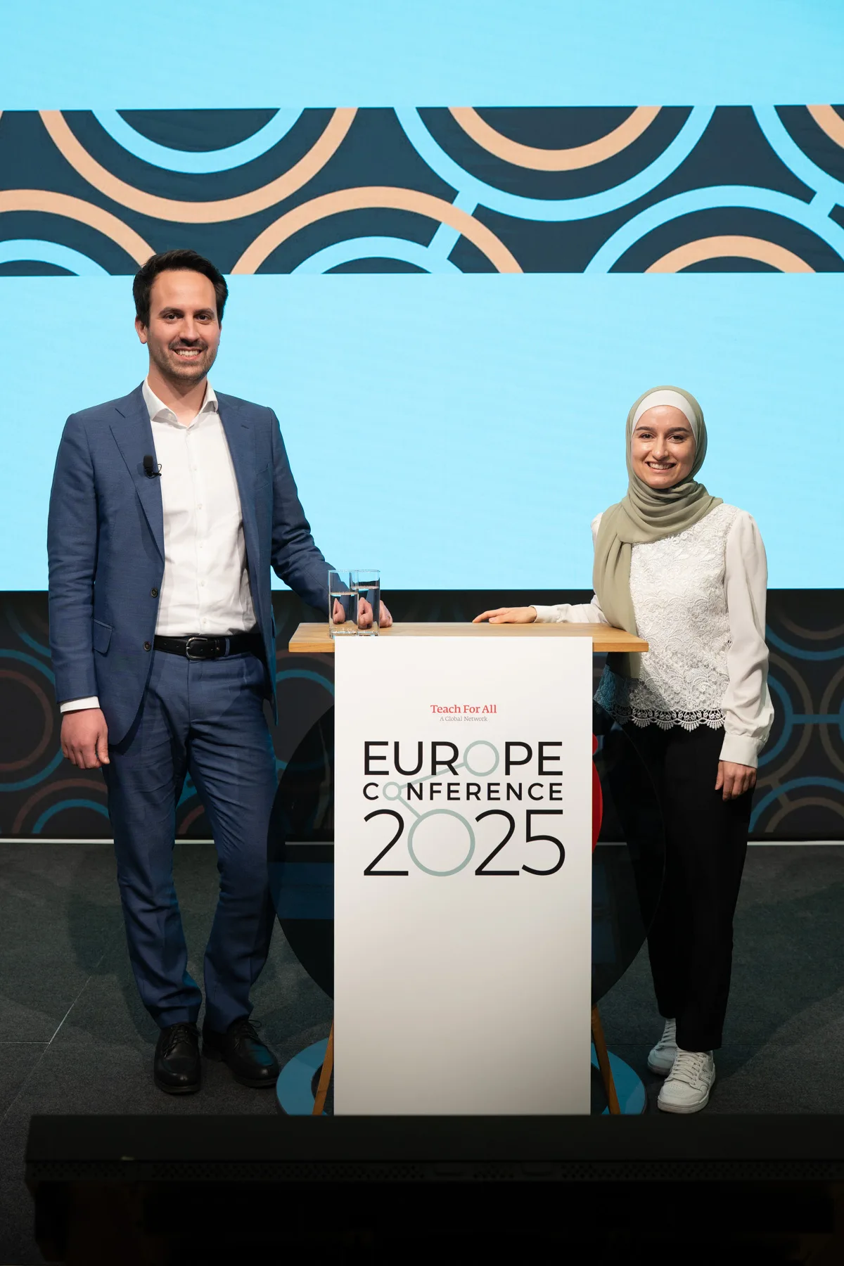 Zwei Personen hinter einem Rednerpult auf einer Bühne der EUROPE CONFERENCE 2025; linke Person im Anzug, rechte Person mit Kopftuch; blaues Kreis-Muster im Hintergrund.