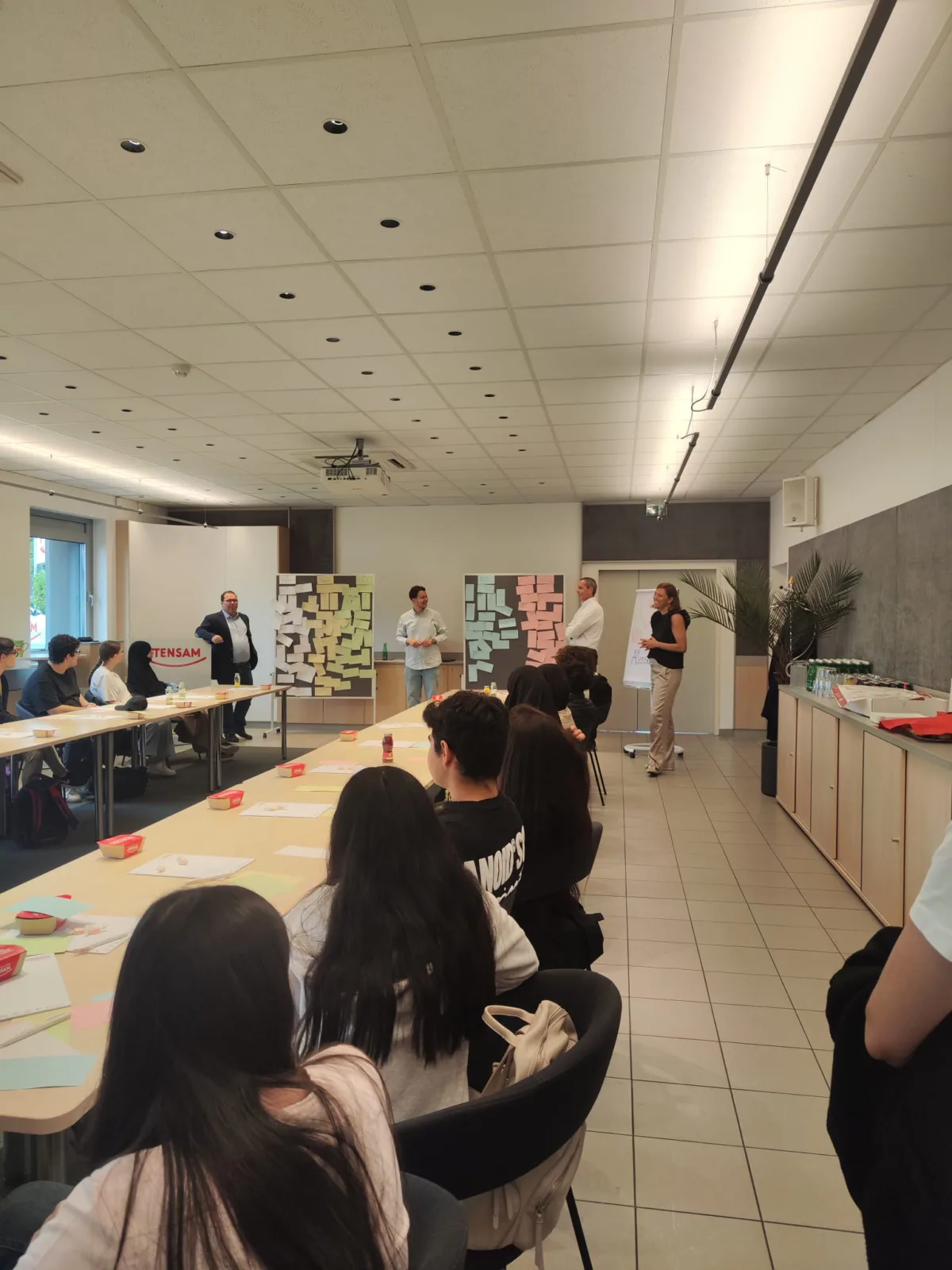 Gruppe von Studierenden in Seminarraum sitzt an langen Tischen; drei Referenten stehen vorne, Pinnwände mit bunten Haftnotizen im Hintergrund.