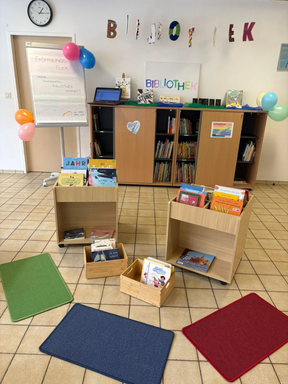 Kinderbibliotheksecke mit Holzregalen voller Bücher, mobilen Buchkisten, bunten Teppichen und Ballons; Bibliothek im Hintergrund.