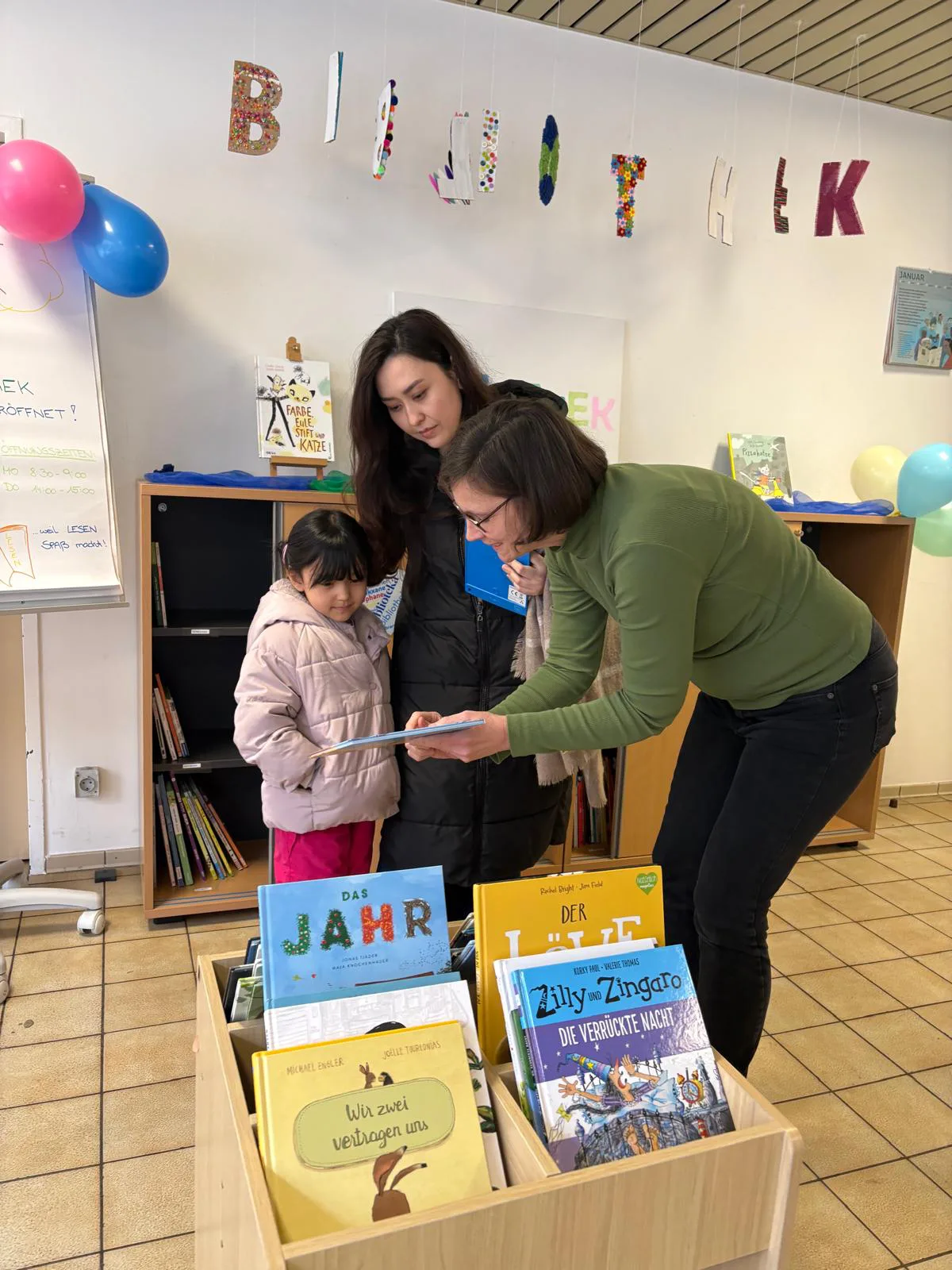 Zwei erwachsene Personen beugen sich über ein Kind in einer Bibliothek mit Kinderbüchern.