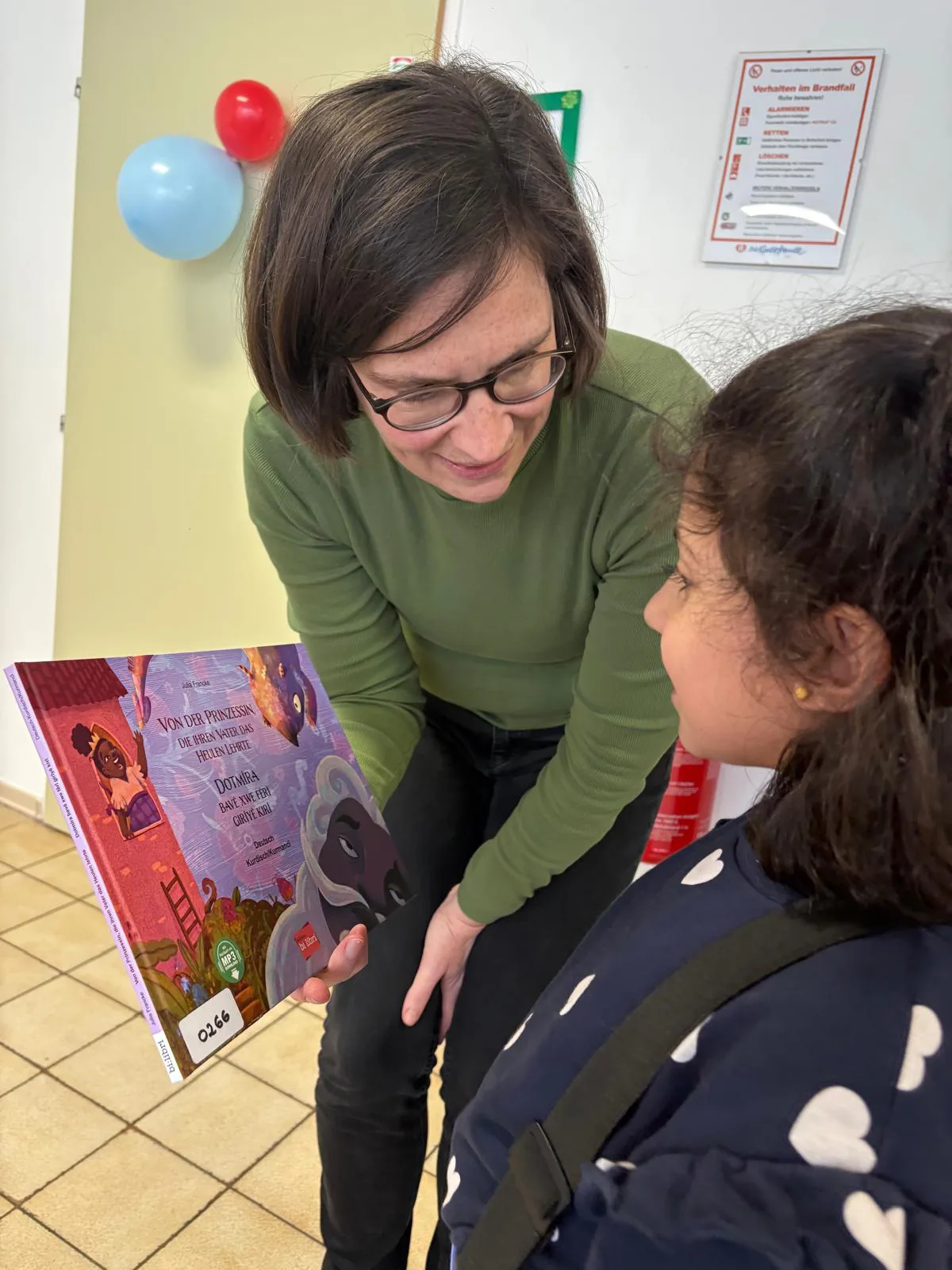 Frau mit Brille beugt sich zu einem Mädchen und zeigt ein buntes Kinderbuch; Schulflur mit Ballons.