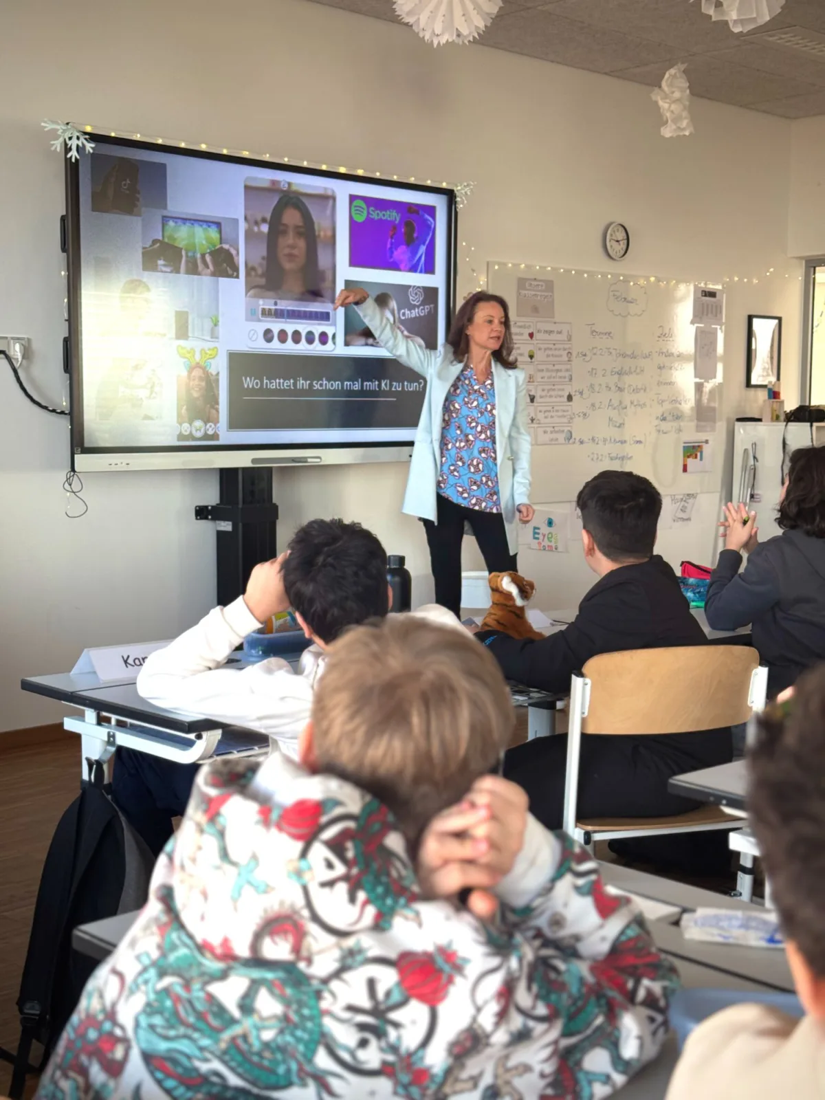 Lehrerin präsentiert Unterricht auf interaktivem Whiteboard in Klassenraum; Lernende sitzen an Tischen und schauen.