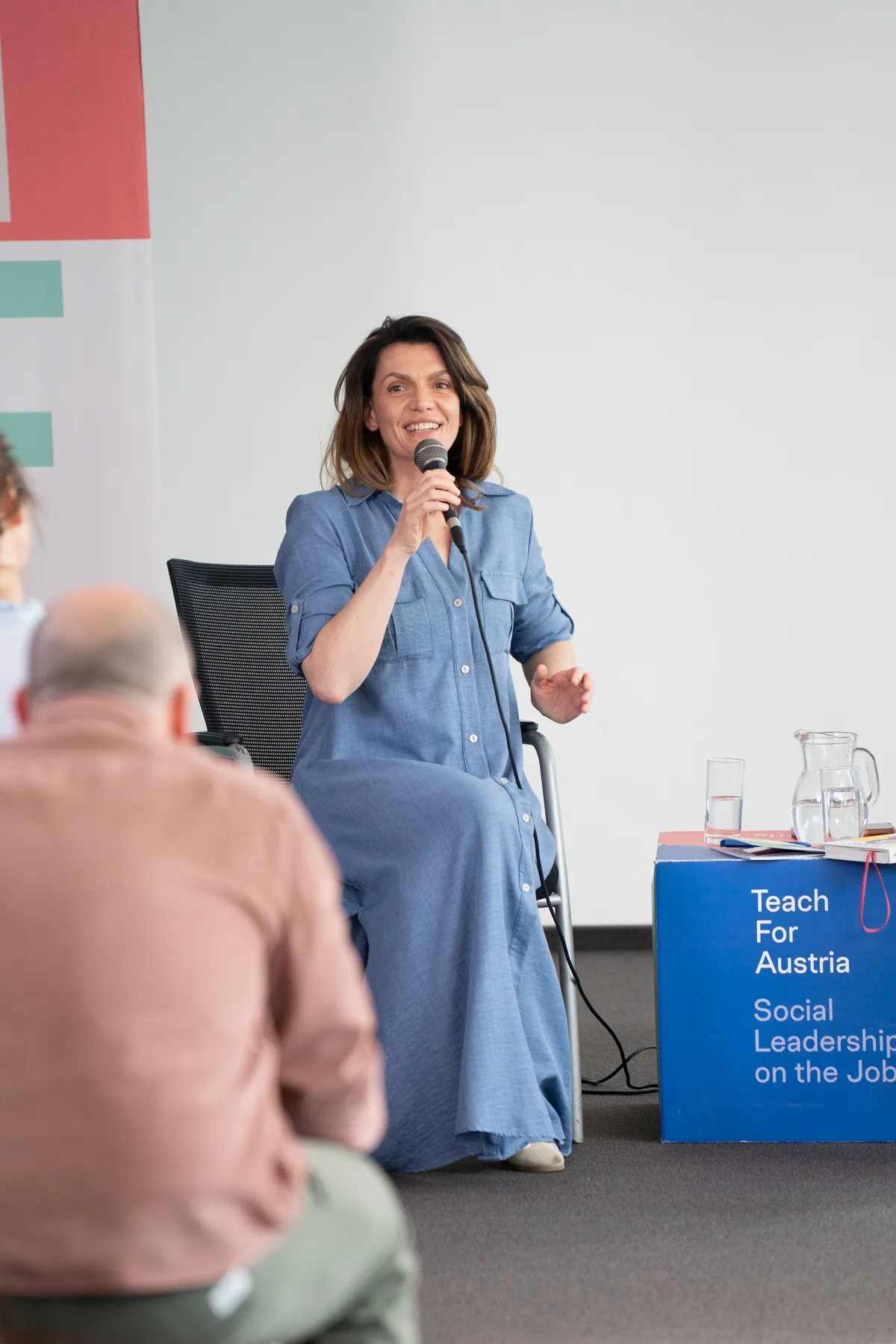 Person in hellblauem Kleid sitzt auf Stuhl, spricht ins Mikrofon bei Konferenz; Tisch mit Gläsern, Text Teach For Austria sichtbar.