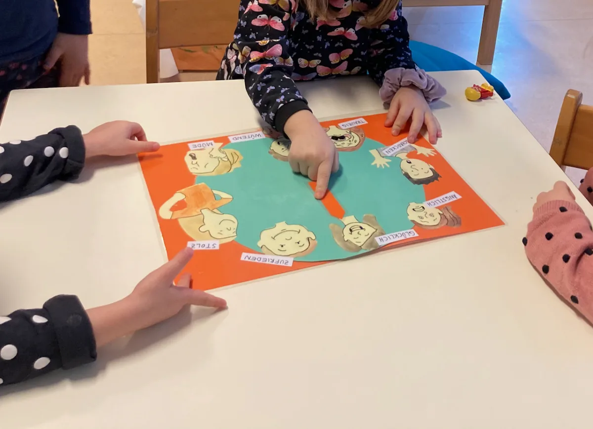 Vier Kinder spielen ein buntes Brettspiel an einem Tisch; eine Hand zeigt auf das Spielbrett.