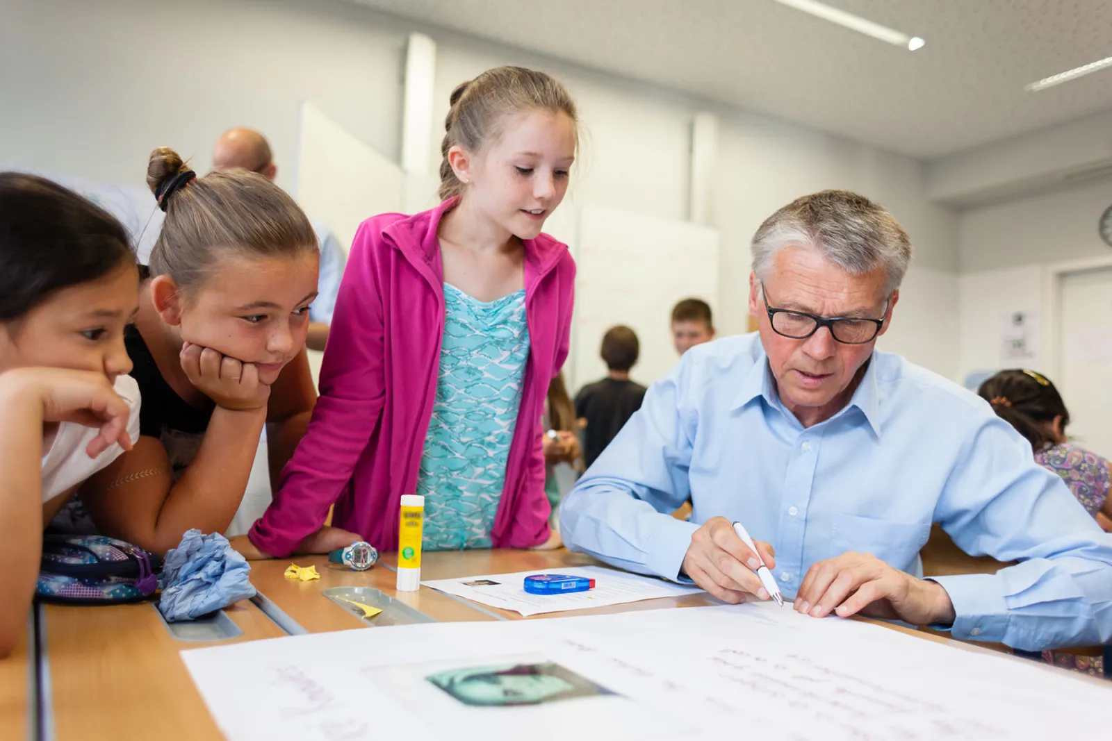 Lehrkraft schreibt auf großem Blatt Papier, während mehrere Schülerinnen im Klassenraum aufmerksam zuschauen.