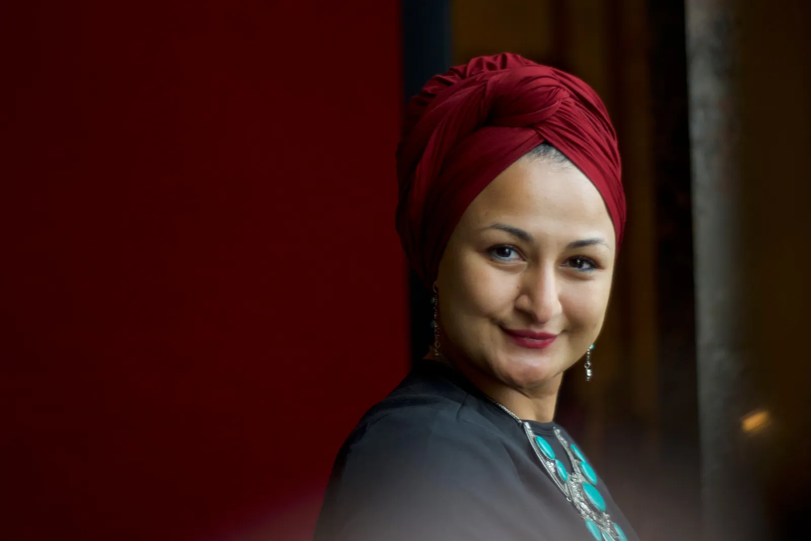 Porträt einer Person mit rotem Turban, lächelt leicht, dunkles Oberteil, türkisfarbener Schmuck, warmes Innenlicht.