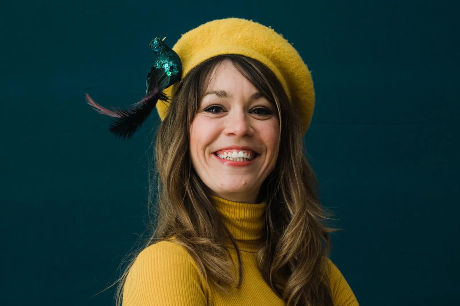 Frau mit gelbem Beret und Feder, gelbem Rollkragenpullover, lächelnd vor dunkelgrünem Hintergrund.