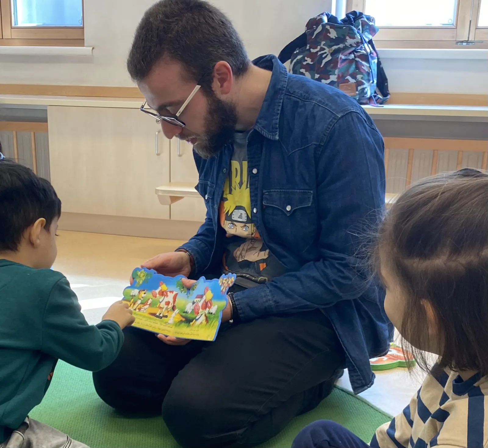 Eine erwachsene Person mit Bart sitzt auf grüner Matte und liest aus einem bunten Bilderbuch für zwei Kinder in einem Klassenzimmer.
