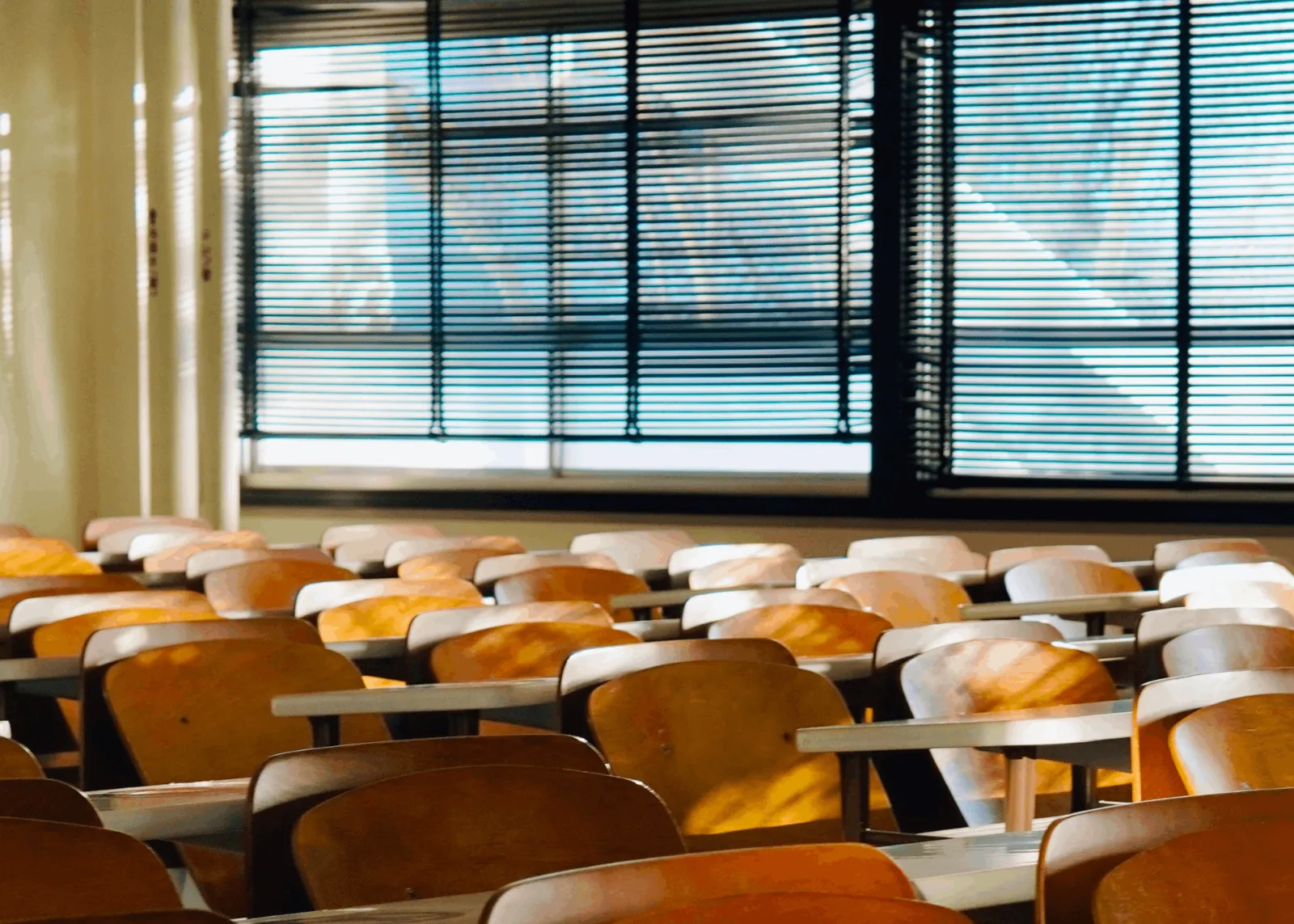 Leeres Klassenzimmer mit Holzstühlen und Tischen; Fenster mit Jalousien, Sonnenlicht fällt hinein.