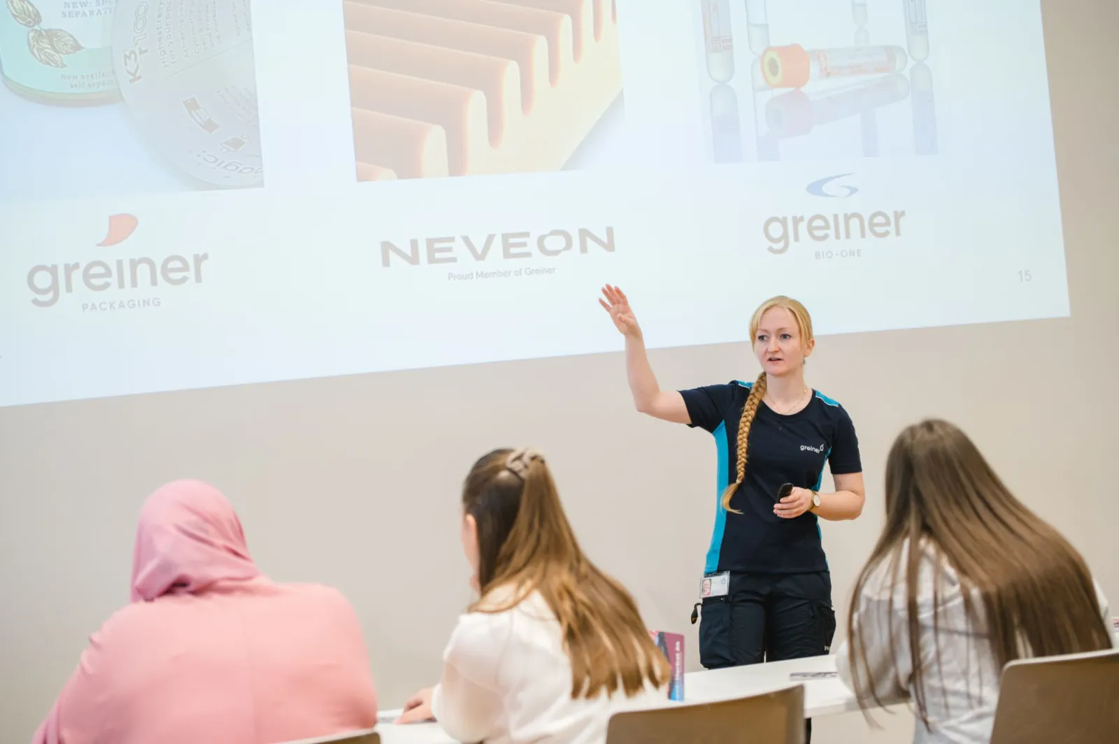 Präsentatorin erklärt vor Publikum, hebt Hand, hält Mikrofon; Projektion zeigt Markenlogos greiner packaging, NEVEON, greiner BIO-ONE.