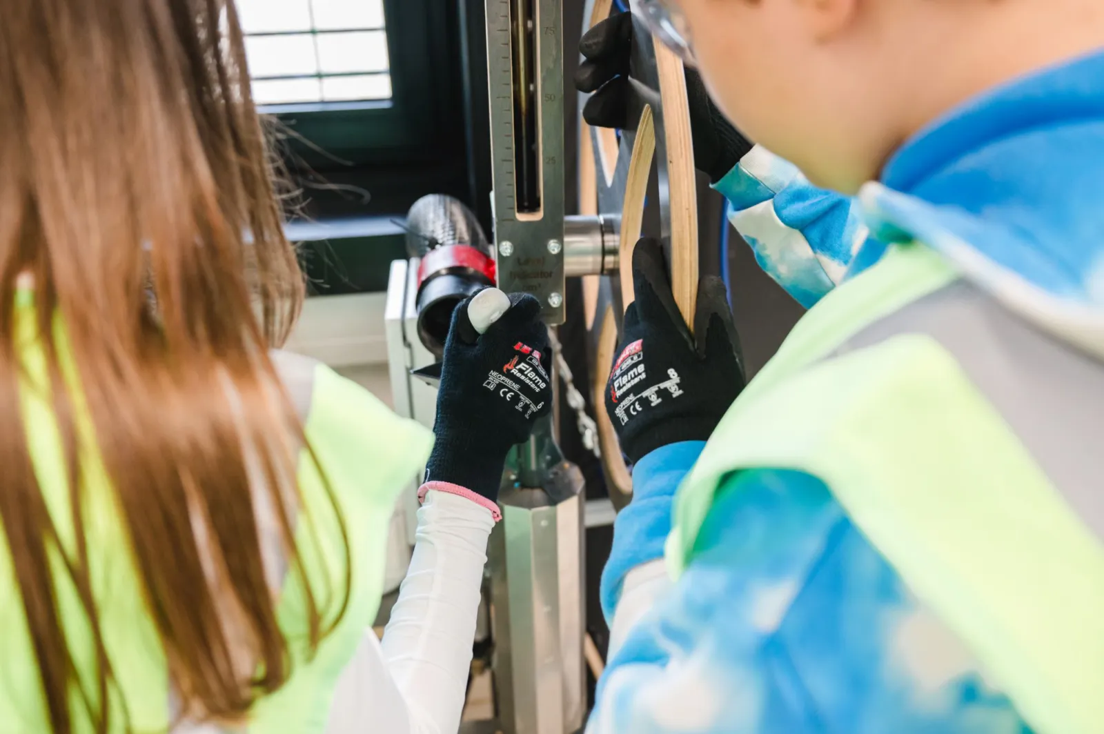 Zwei Fachkräfte in Schutzkleidung justieren eine Messvorrichtung an einer Metallstruktur in einer Werkstatt.