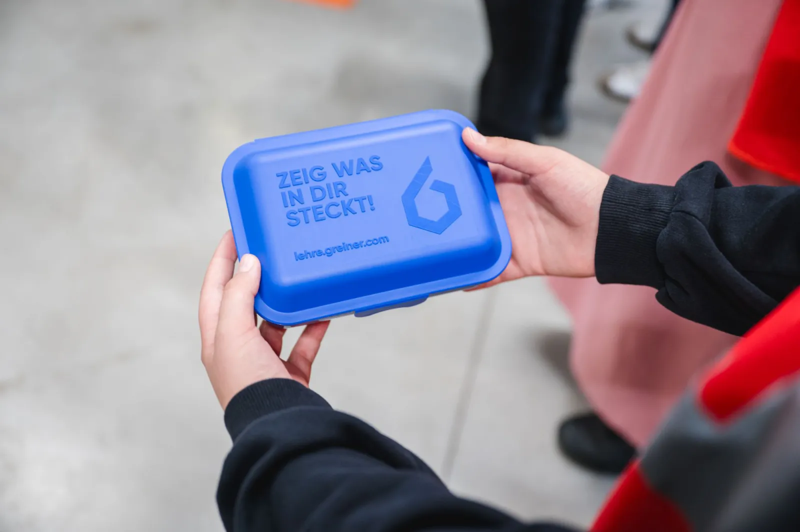 Zwei Hände halten eine blaue Lunchbox mit dem Aufdruck 'ZEIG WAS IN DIR STECKT!' bei einer Veranstaltung.