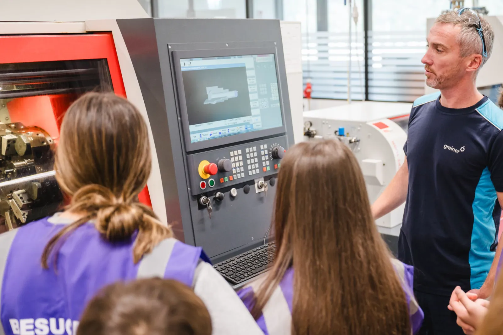 Auf einer CNC-Fräsmaschine erklärt ein Mann zwei Besucherinnen das Bedienfeld der Maschine in einer Werkstatt.