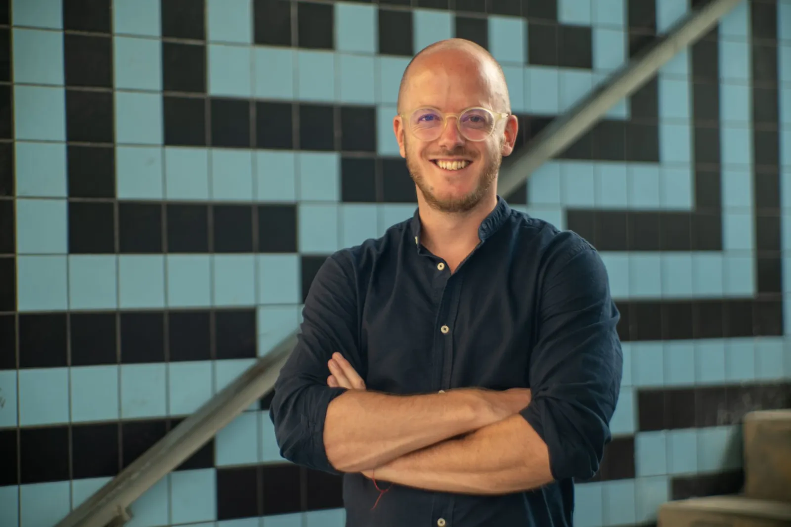 Person mit Brille und Bart steht mit verschränkten Armen vor gemusterter Fliesenwand neben Treppe.