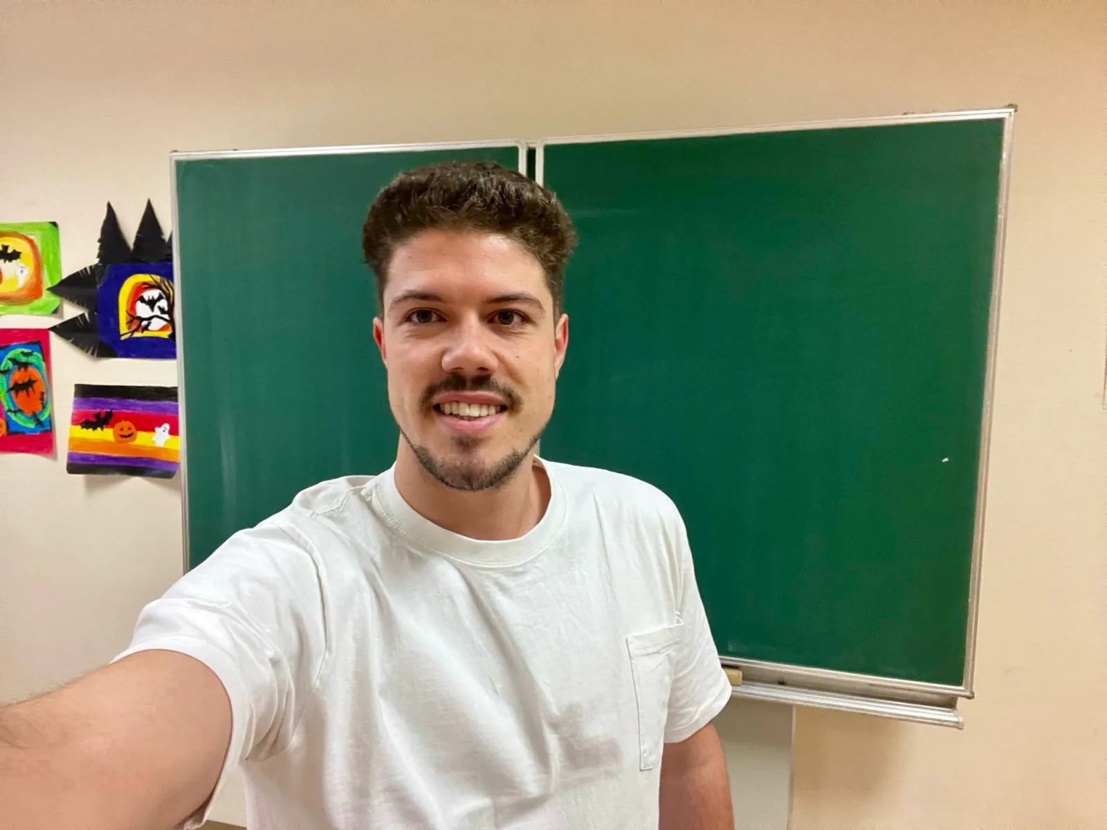 Person macht Selfie vor grünem Kreidetafel im Klassenzimmer; trägt weißes T-Shirt; linke Wand mit bunten Kunstwerken.