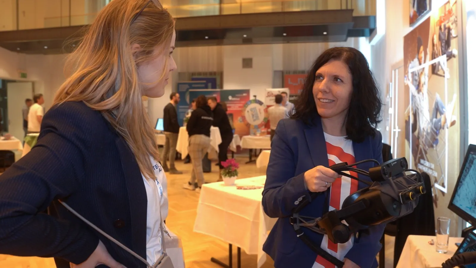 Zwei Frauen auf einer Messe; eine präsentiert der anderen ein VR-Headset, während beide lächeln; Messestände und Banner im Hintergrund.