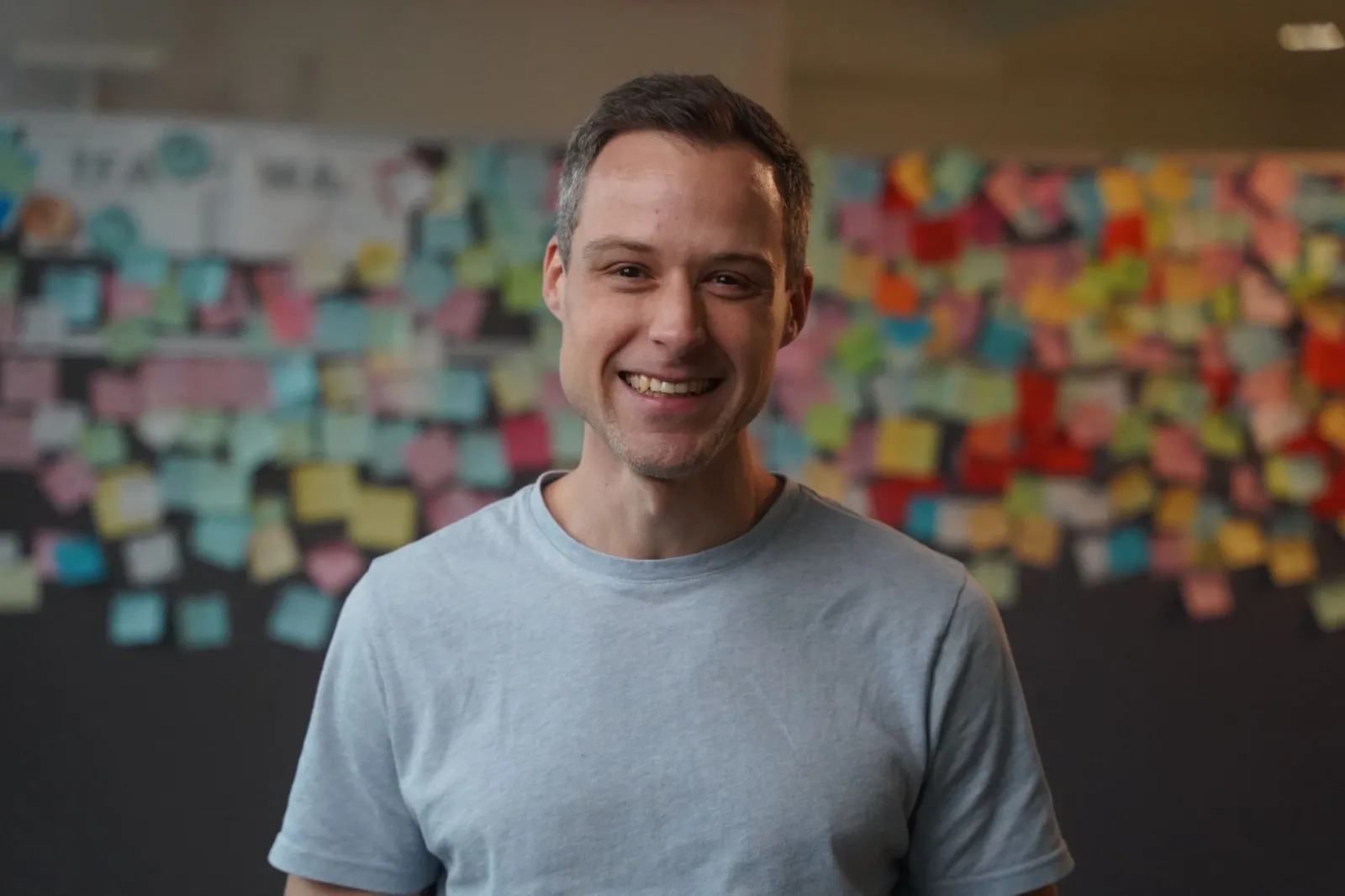 Person lächelt in einem Büro vor Wand mit bunten Post-it-Notizen.