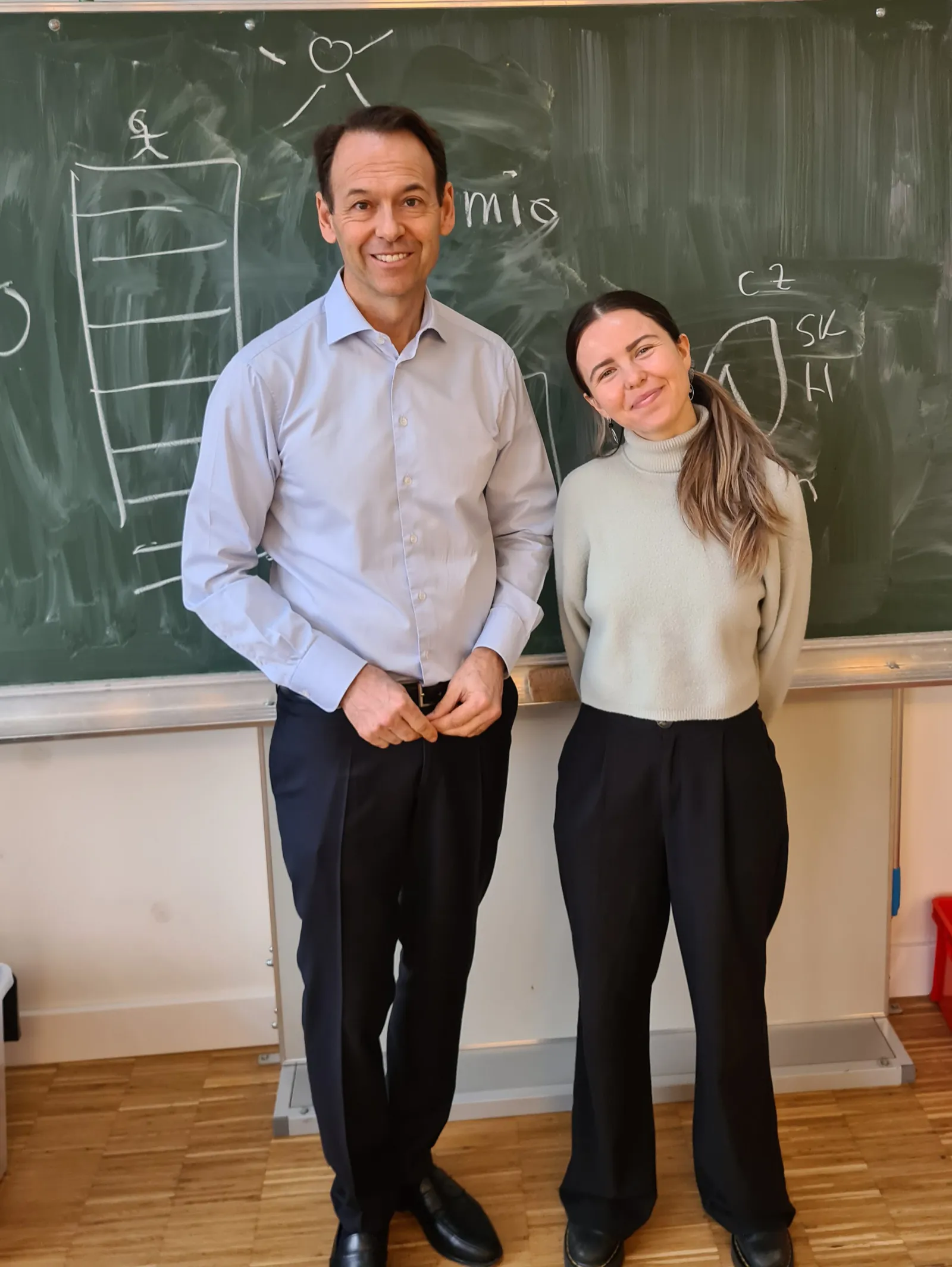 Zwei Personen posieren lächelnd vor einer grünen Tafel in einem Klassenzimmer.