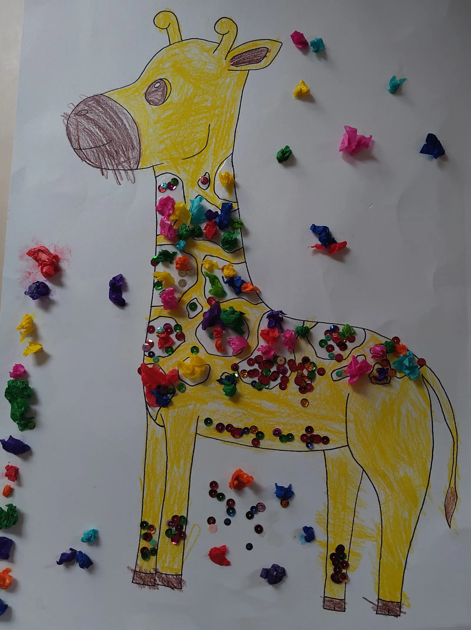 Gelbe Giraffe steht auf weißem Blatt; Körper mit bunten Perlen verziert, buntes Konfetti um sie herum.
