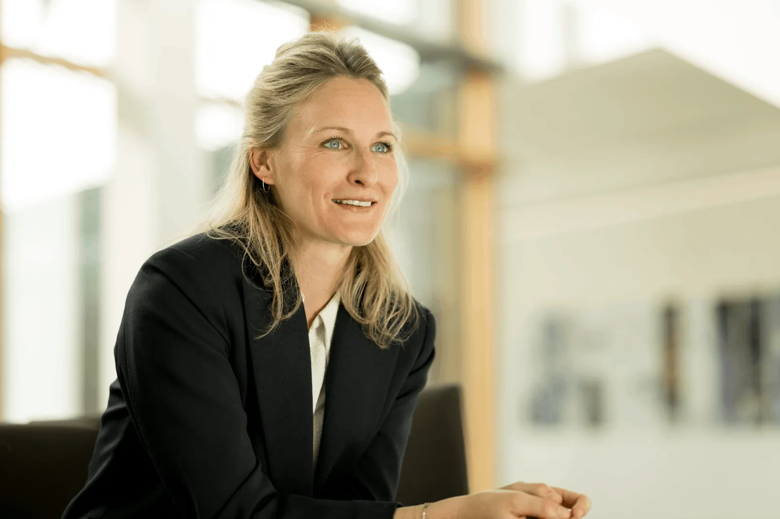 Geschäftsperson in schwarzem Blazer lächelt im hellen Büro, sitzt am Tisch.