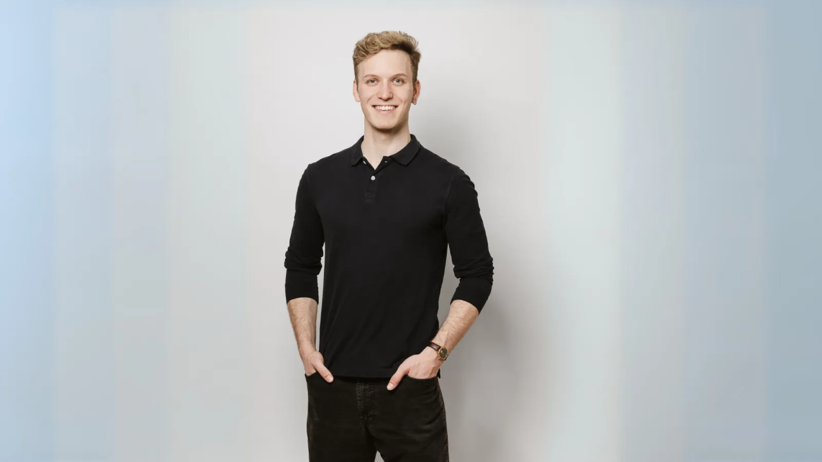 Junger Mann in schwarzem Polohemd steht frontal, Hände in den Hosentaschen, lächelnd vor hellem Studiohintergrund.