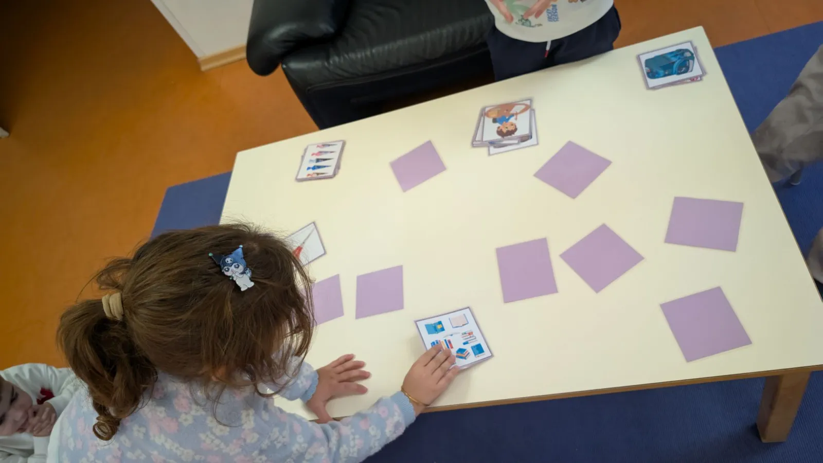 Kind greift Karte vom Memory-Spieltisch; violette Quadrate liegen verstreut, weitere Spielkarten sichtbar.