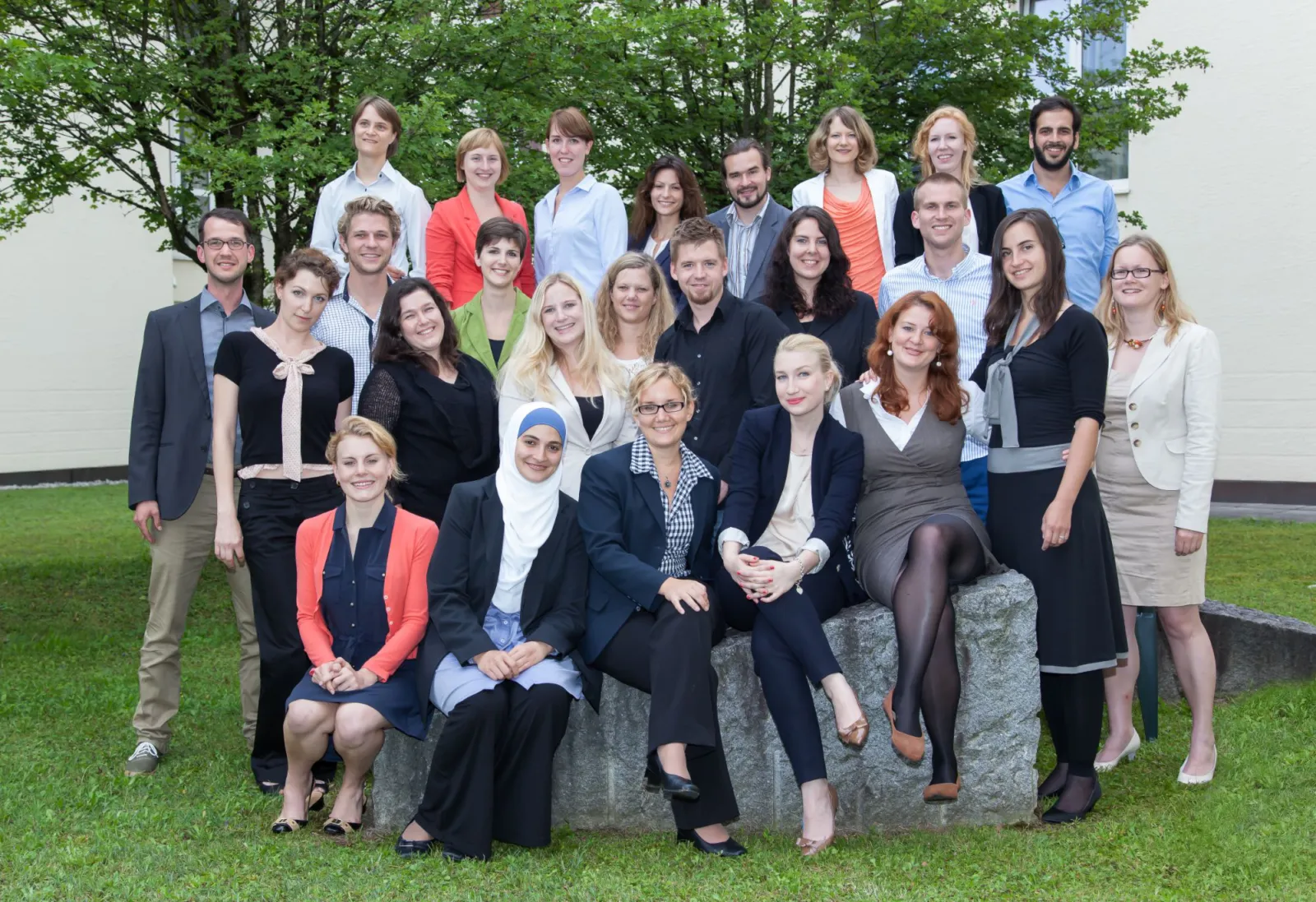 Gruppenporträt von Erwachsenen, die im Freien posieren; sie stehen und sitzen auf einem Steinblock vor Gras, Bäumen und einem Gebäude.