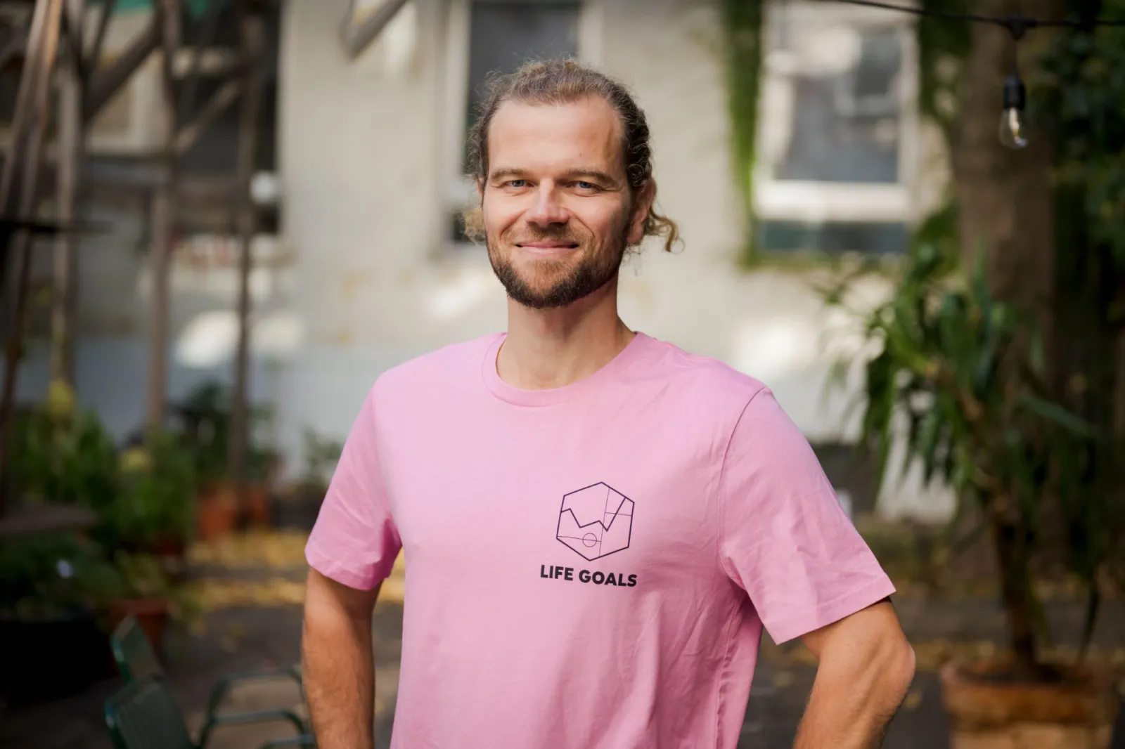 Person in rosa T-Shirt mit Logo Life Goals steht lächelnd im Garten, Hände an den Hüften.