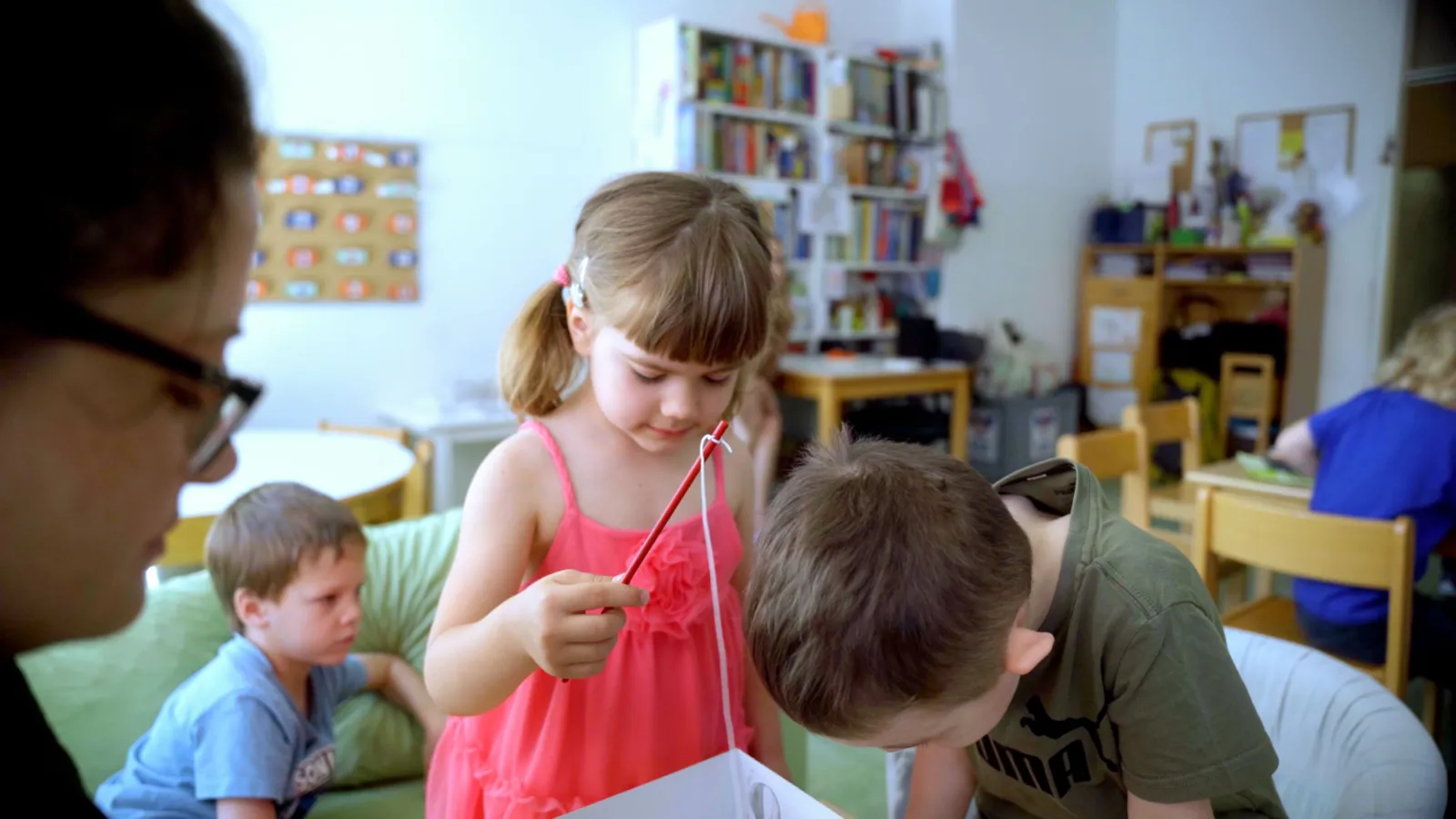 Kind im pinken Kleid bastelt; hält rotes Bastelwerkzeug mit weißem Faden im Klassenraum.
