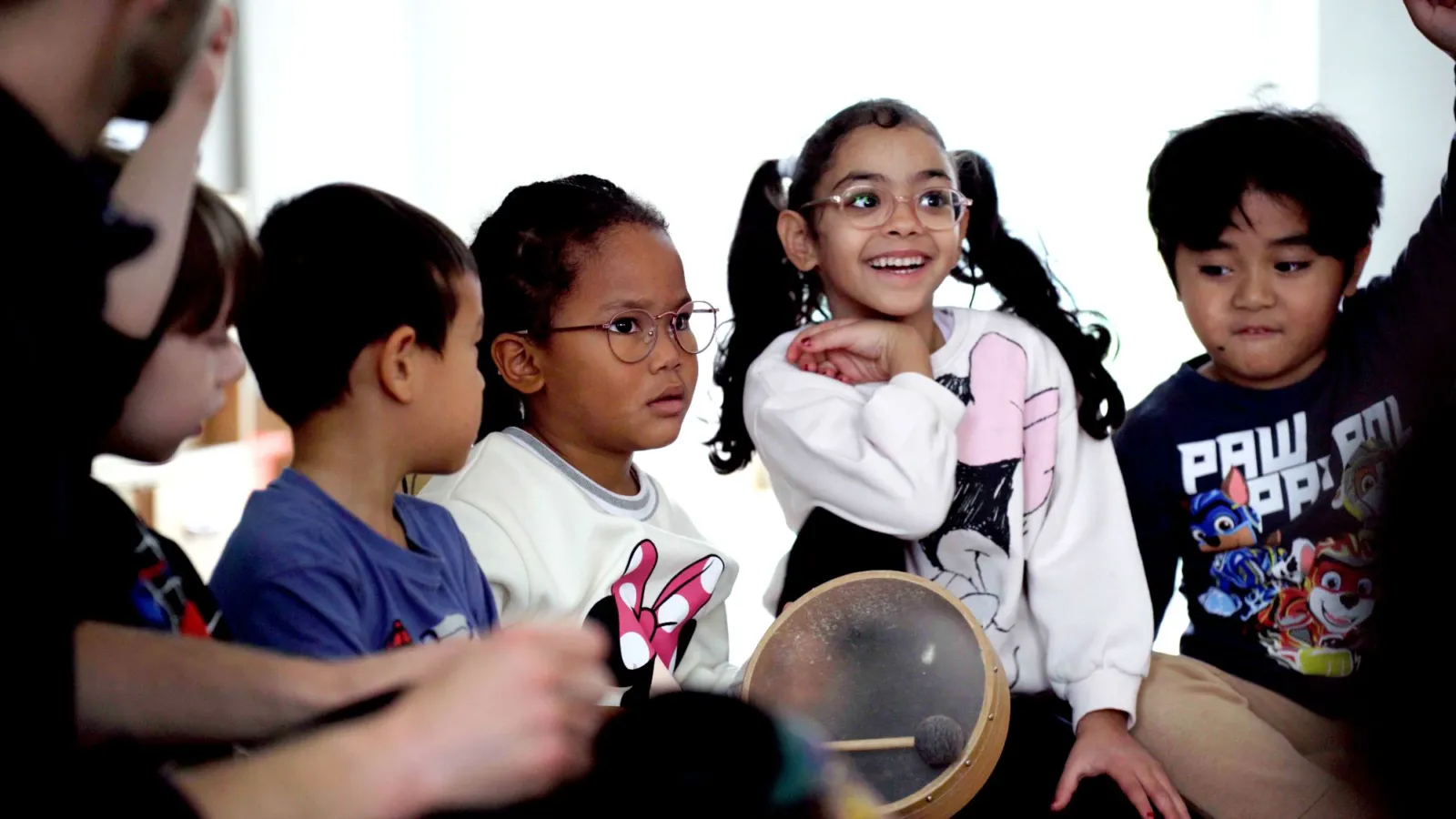 Gruppe von Kindern sitzt im Kreis, zentrale Rahmentrommel vor ihnen, beim Musikunterricht.