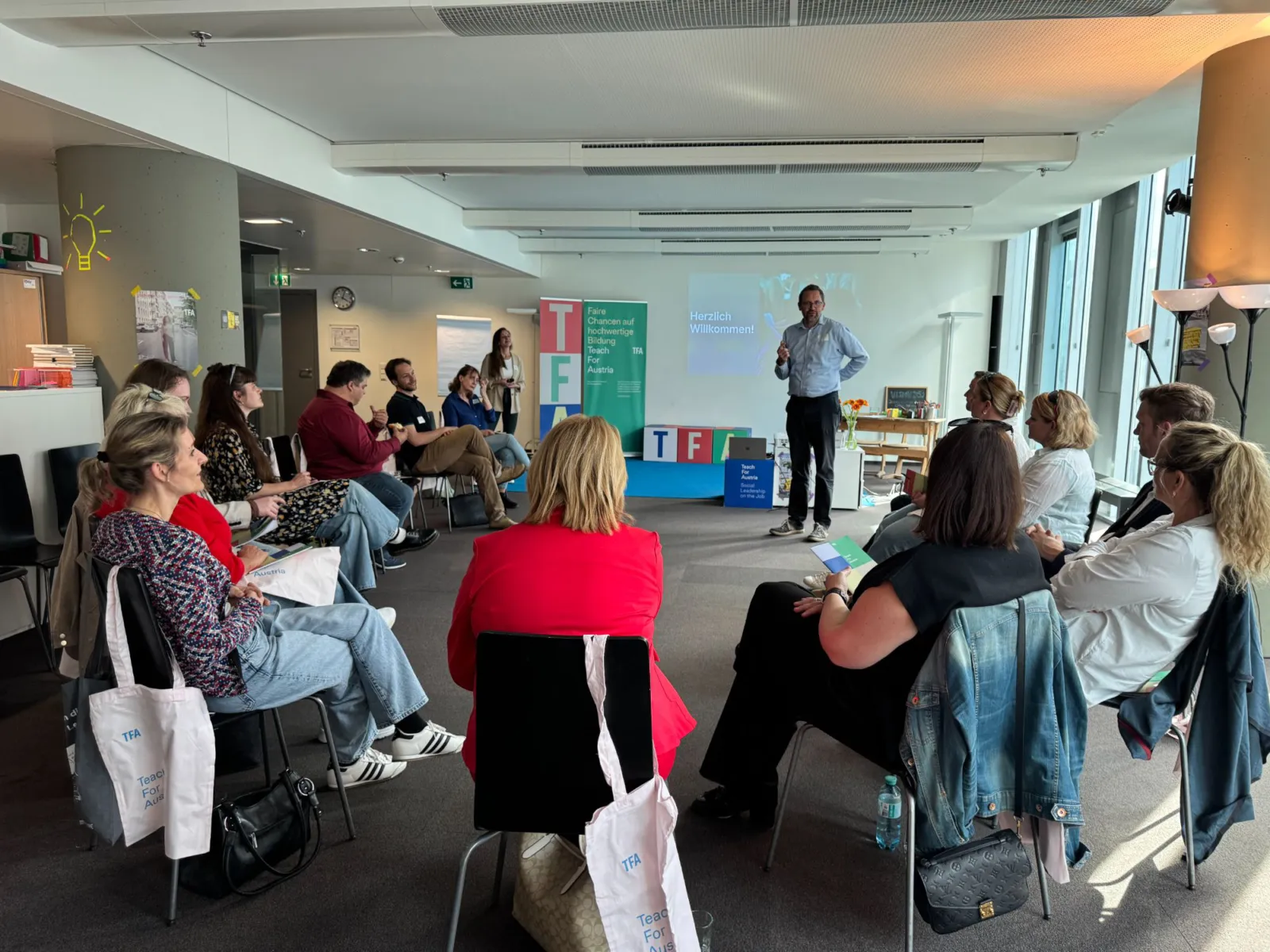 Sprechende Person steht vorne und spricht vor Gruppe in Konferenzraum; Projektion 'Herzlich Willkommen' im Hintergrund.