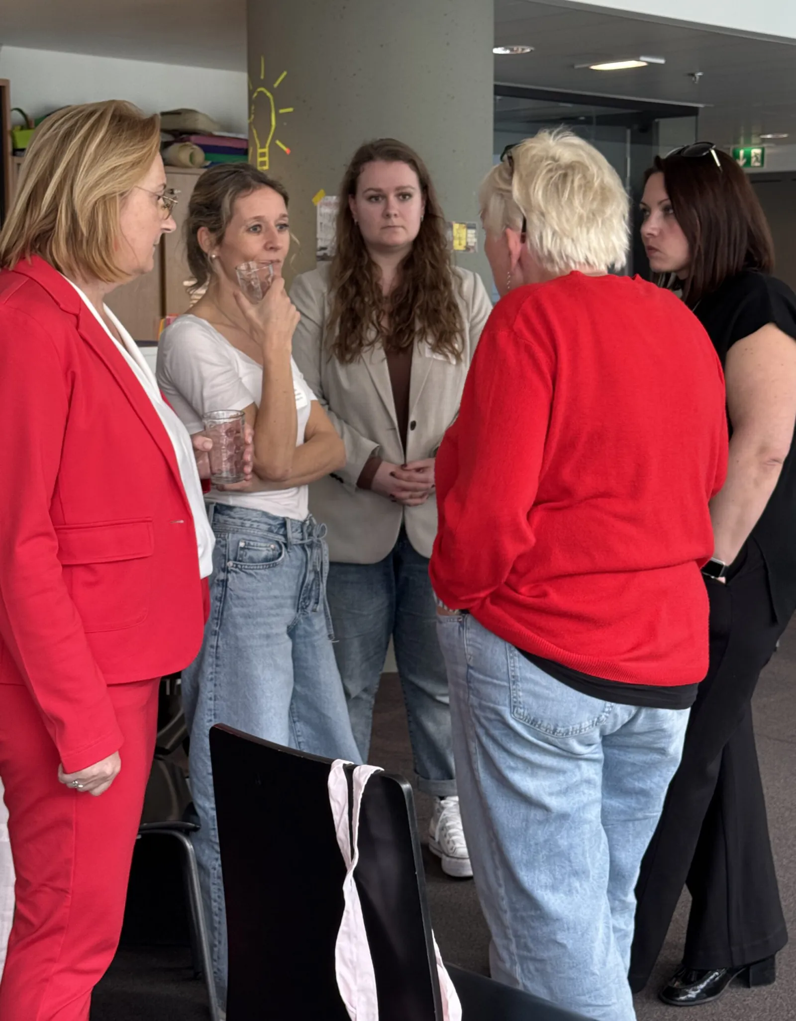 Fünf Erwachsene stehen im Kreis und unterhalten sich; zwei halten Gläser, Hintergrund wirkt wie Bürobereich mit Säule.