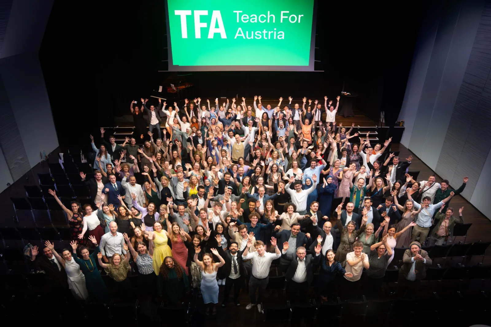 Große Gruppe von Menschen auf Bühne, Arme erhoben, vor grünem Bildschirm mit 'TFA Teach For Austria'.