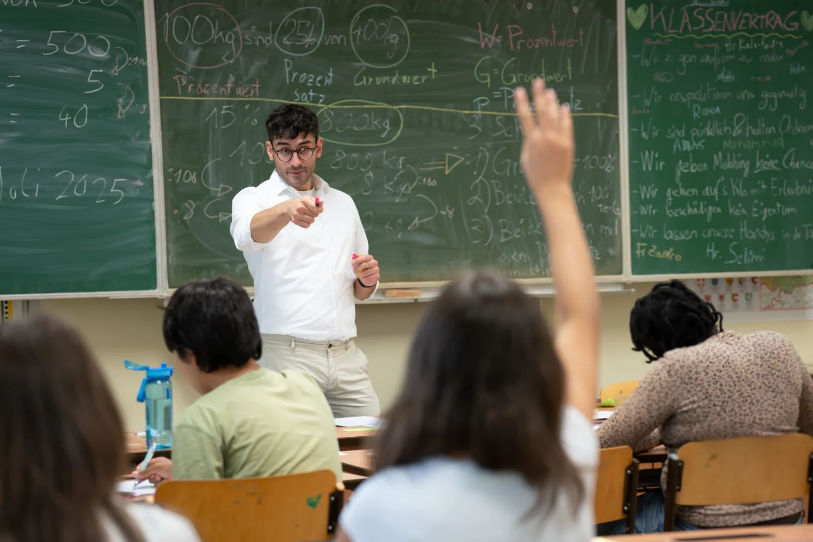 Lehrkraft erklärt im Klassenzimmer vor grüner Tafel mit Formeln; mehrere Lernende sitzen, einer hebt die Hand.