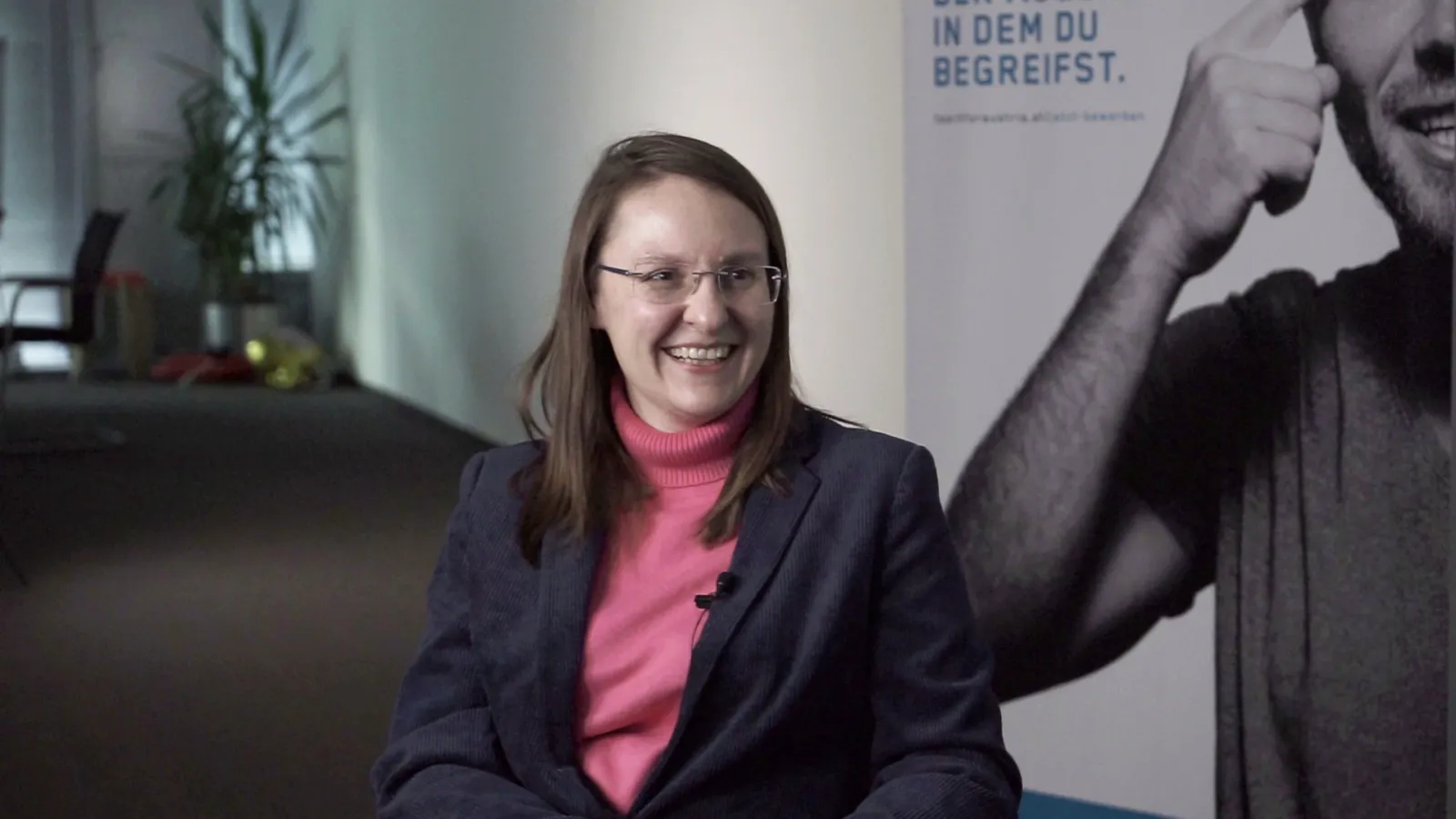 Person sitzt, lächelt, in dunkelblauem Blazer und pinkem Rollkragenpullover; Ansteckmikrofon sichtbar, Hintergrund mit Schwarz-Weiß-Poster.