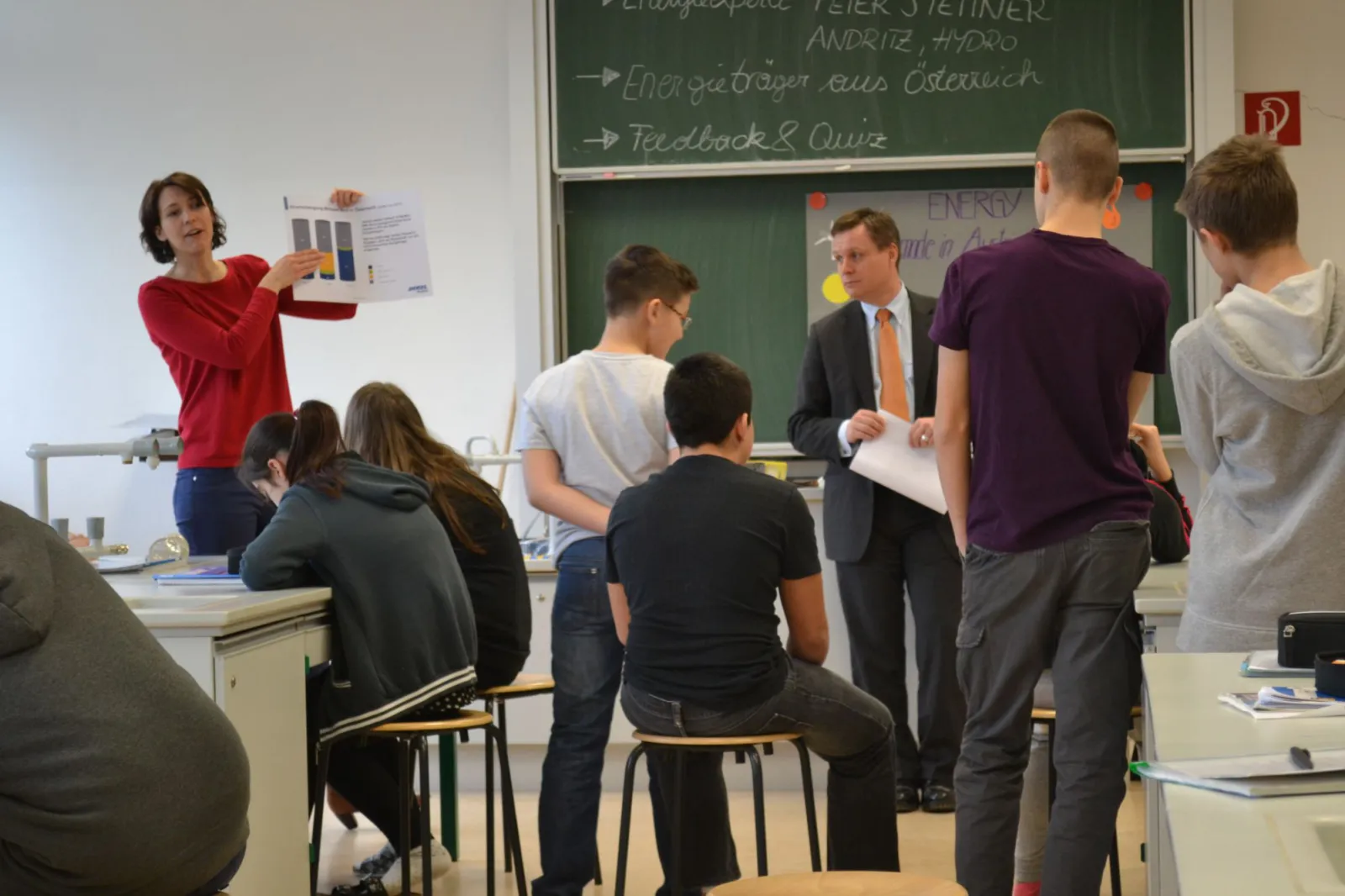 Lehrkraft präsentiert Diagramm-Poster vor Lernenden in einem Klassenraum.