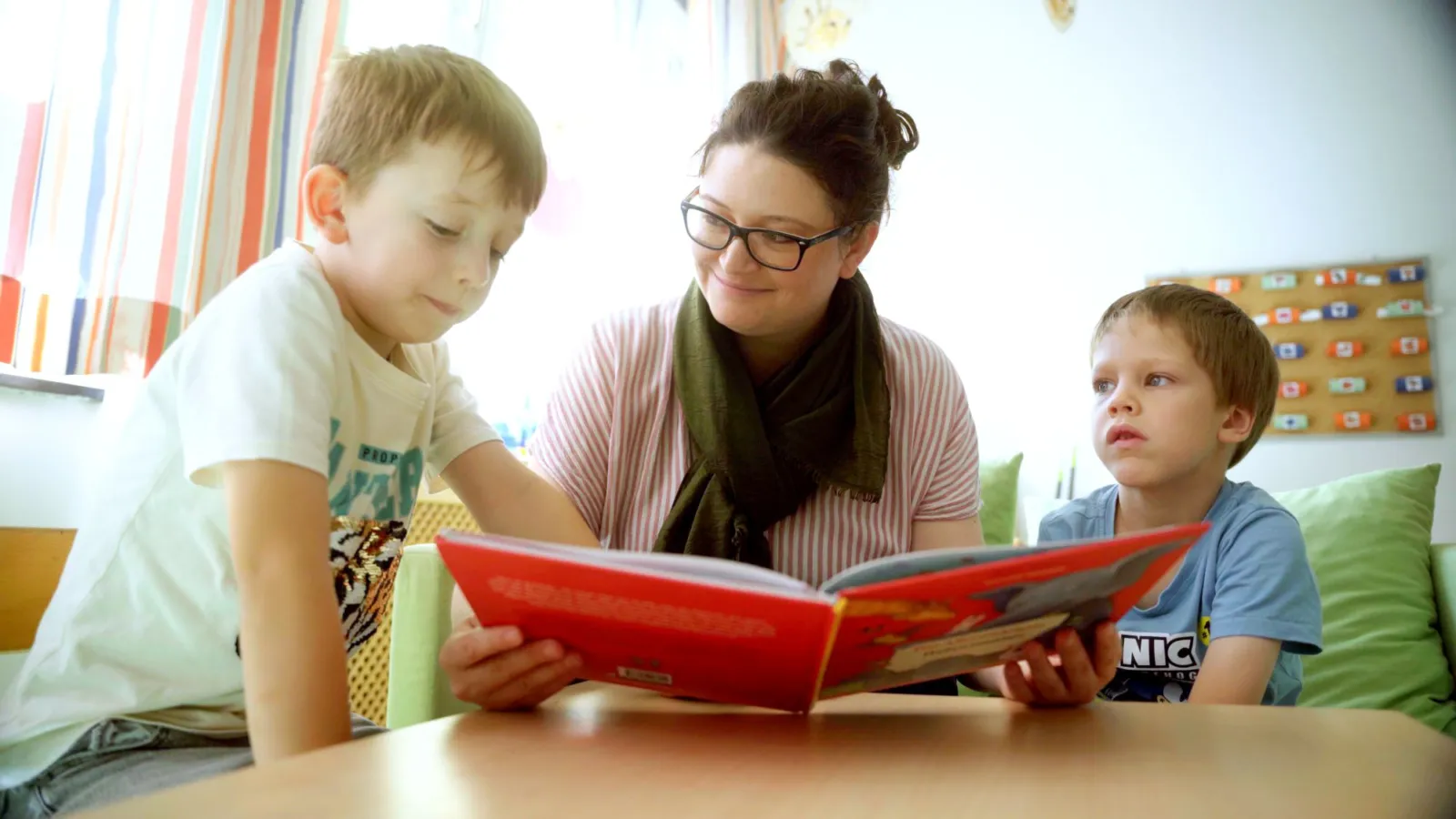 Erzieherin liest zwei Jungen in hellem Kita-Raum aus rotem Buch vor.