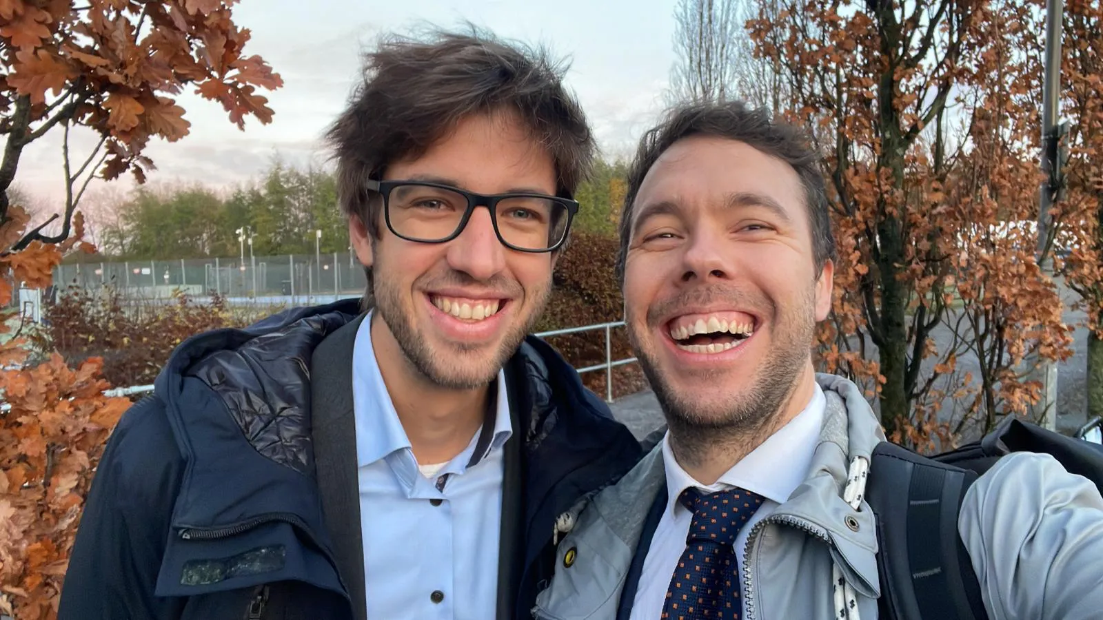 Zwei Männer machen ein Selfie im Freien, lächelnd vor Herbstlaub an Bäumen; Geländer im Hintergrund.