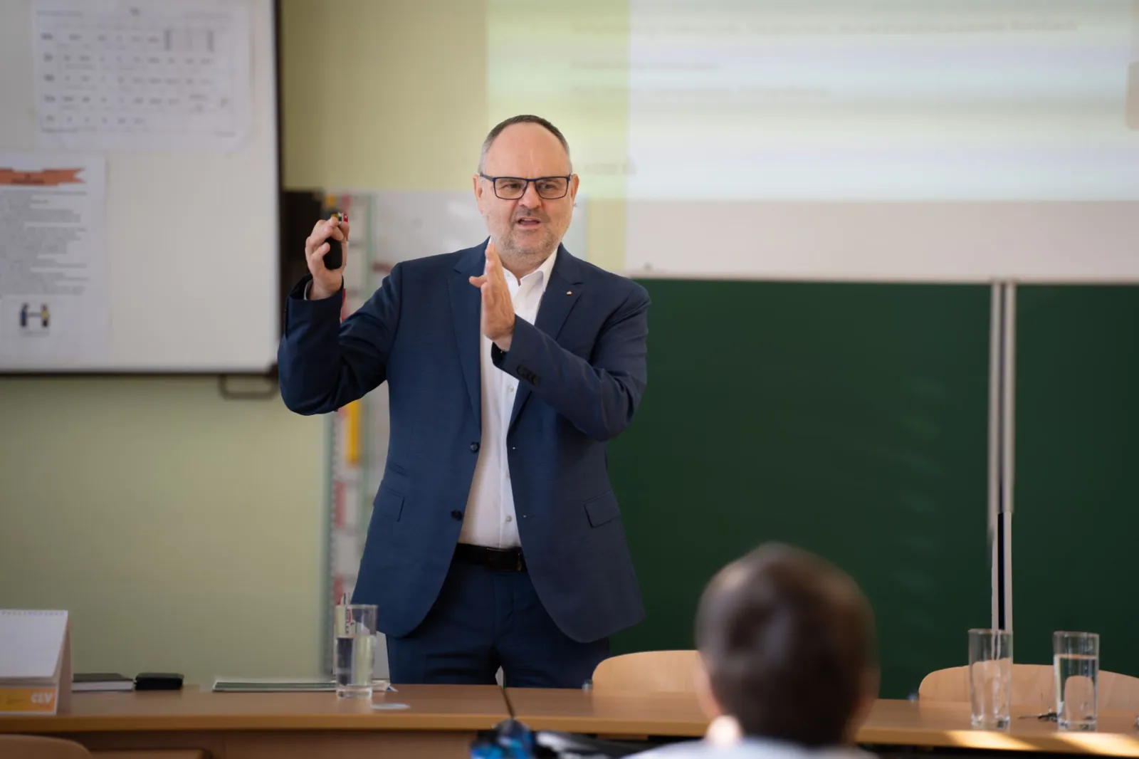 Lehrkraft in dunkelblauem Anzug präsentiert vor Klasse, nutzt Fernbedienung; Hintergrund mit Tafel und Beamer.