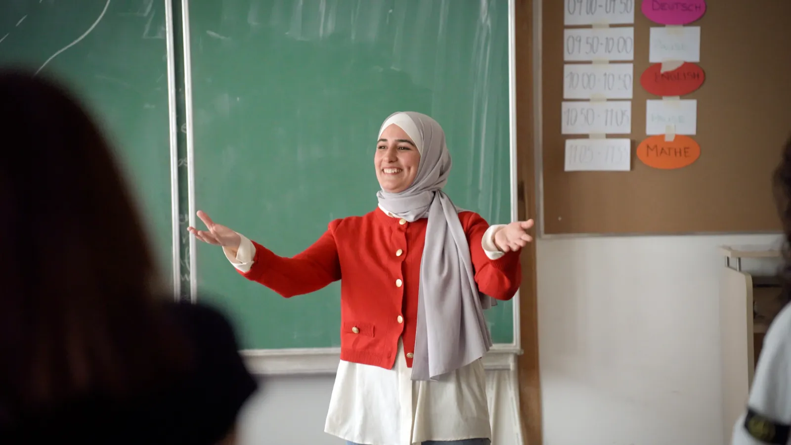 Lehrkraft in roter Jacke vor grüner Tafel, Arme ausgebreitet, spricht zu Lernenden im Klassenzimmer.