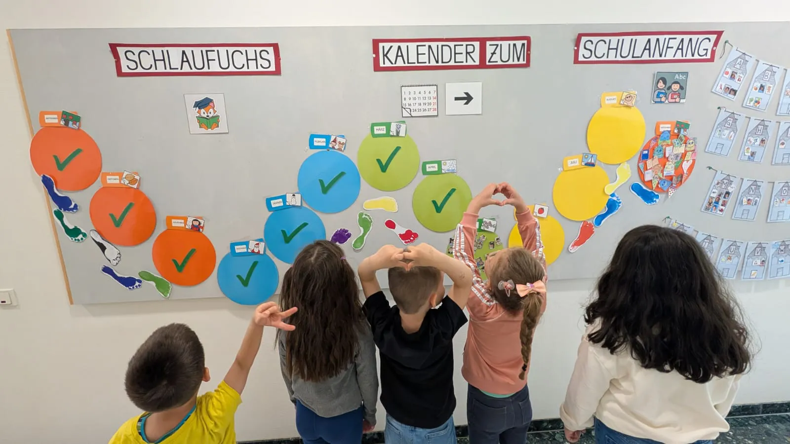 Gruppe von Kindern von hinten vor einer Schulwand mit bunten Kreisen und Häkchen; sie formen ein Herz mit den Armen.
